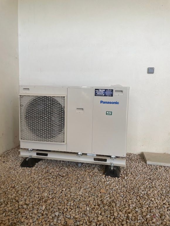 Une unité de pompe à chaleur Panasonic montée sur des blocs, posés sur du gravier, contre un mur.