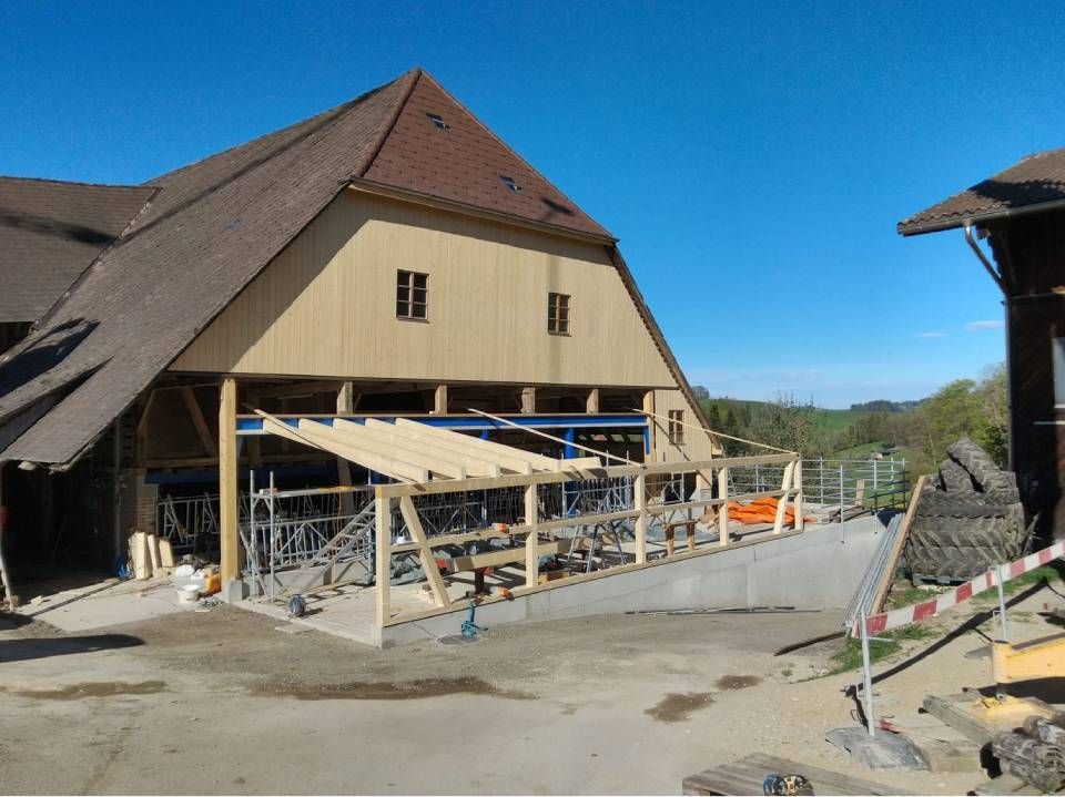 Baustelle mit einem scheunenartigen Gebäude, Gerüsten und Betonarbeiten unter klarem, blauem Himmel