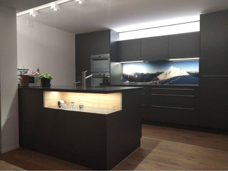 Moderne Küche in Schwarz und Holz mit zentraler Kochinsel und beleuchtetem Fliesenspiegel sowie Schränken mit Berglandschaftsmotiv.
