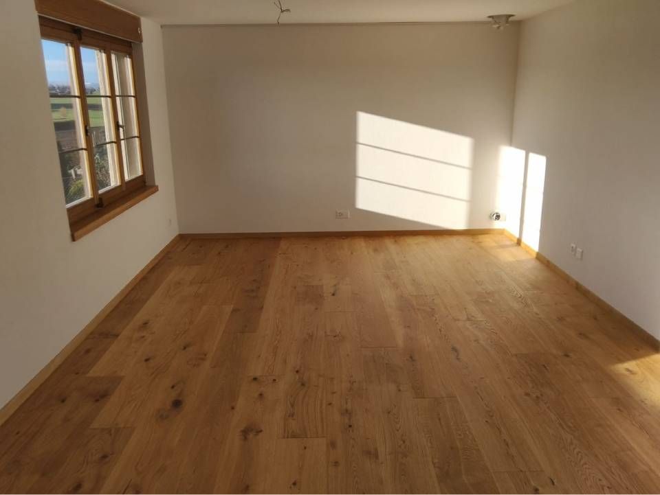 Leerer, sonnendurchfluteter Raum mit Holzboden, weissen Wänden und einem Fenster, das Schatten wirft.