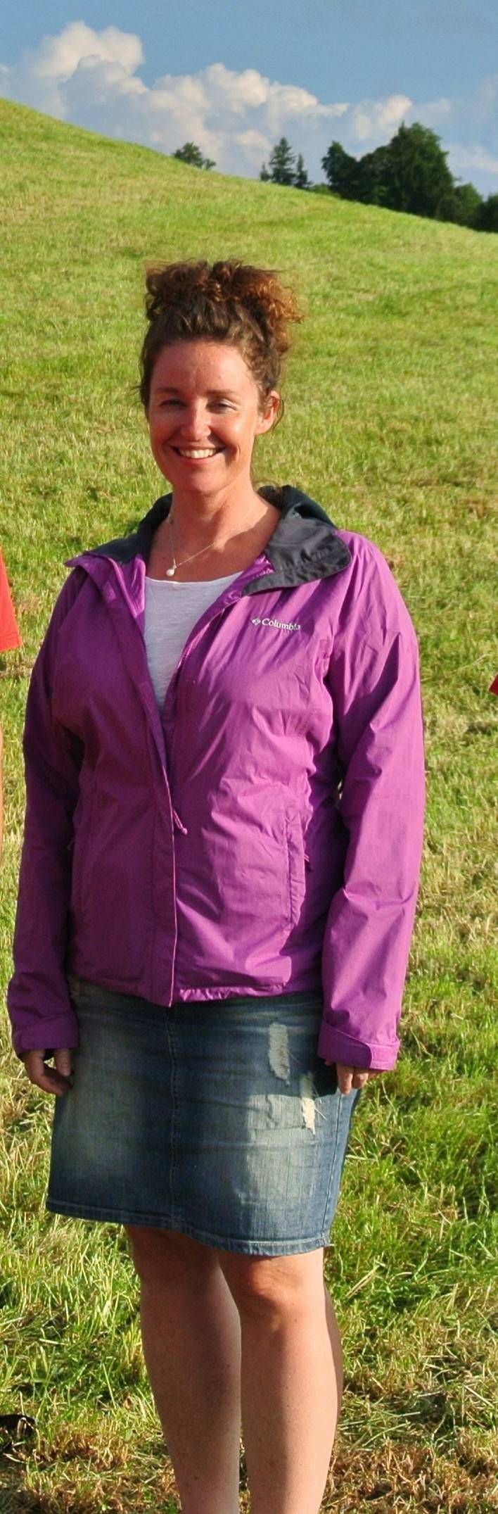 Eine Person steht im Freien auf einem grasbewachsenen Hügel. Sie trägt eine lila Jacke und einen Jeansrock. Im Hintergrund sind blauer Himmel und Wolken zu sehen.