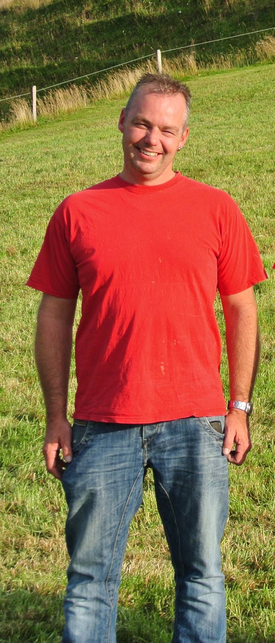 Mann in rotem T-Shirt und Jeans steht lächelnd im Gras und lächelt ins Sonnenlicht.