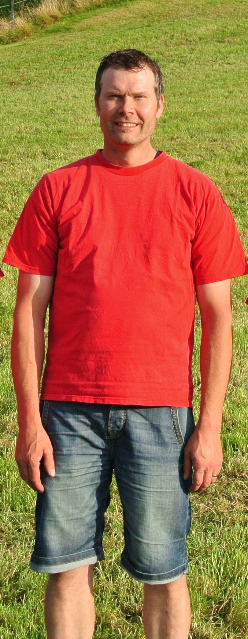 Mann steht im Freien auf einer Wiese und trägt ein rotes T-Shirt und Jeansshorts.
