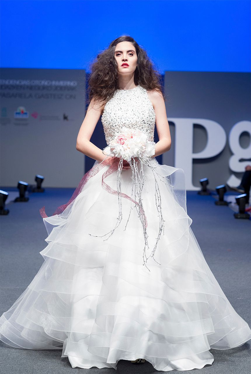 Una mujer con un vestido de novia camina por una pasarela sosteniendo un ramo de flores.