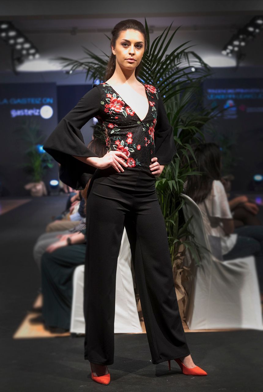 Una mujer camina por una pasarela vistiendo un top floral y pantalones negros.