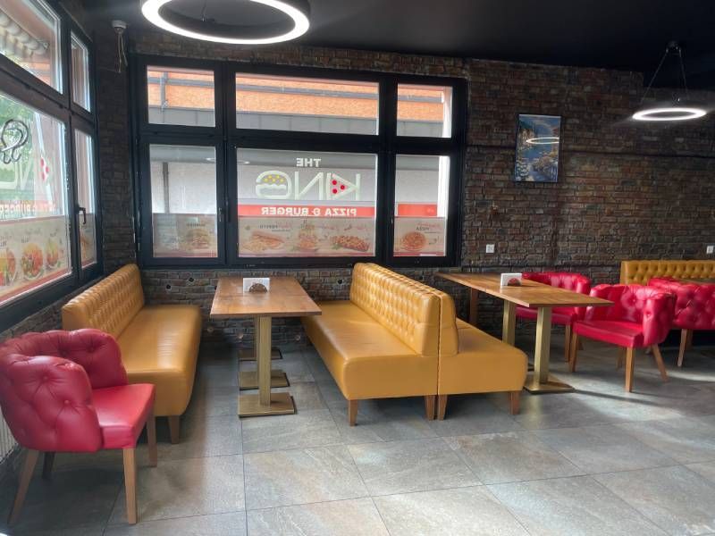 Blick in Gastraum von The King Pizzeria & Burger, links senffarbenes Kunstledersofa mit Holztisch und weiterem Sofa vor Fensterfront, weiteres senffarbendes Sofa mit Holztisch und zwei roten Kunstlederstühlen, rechts angeschnitten ein weiteres Kunstledersofa in Senfgelb und ein roter Kunstlederstuhl, Wand in Ziegeloptik, grau-beiger Fließenboden