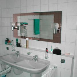 Waschbecken mit Spiegel, Handtüchern und Toilettenartikeln. Weiße Fliesen, brauner Spiegelrahmen und blaugrünes Handtuch.