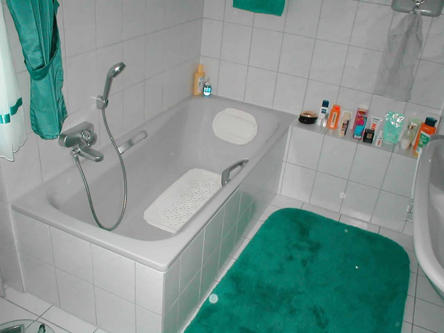 Badezimmer mit weiß gefliesten Wänden, einer grauen Badewanne, einem grünen Teppich und Toilettenartikeln auf einem Sims.