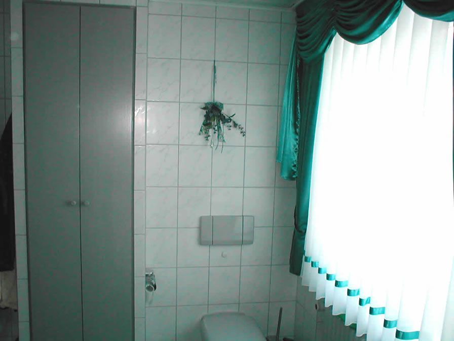 Badezimmer mit einem hohen, grauen Schrank, weiß gefliesten Wänden und einem Fenster mit grünen Vorhängen.