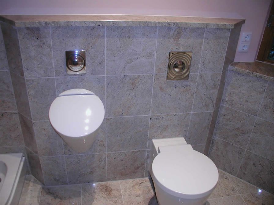 Modernes Badezimmer mit Urinal, Toilette und Spülknöpfen an einer grau gefliesten Wand.