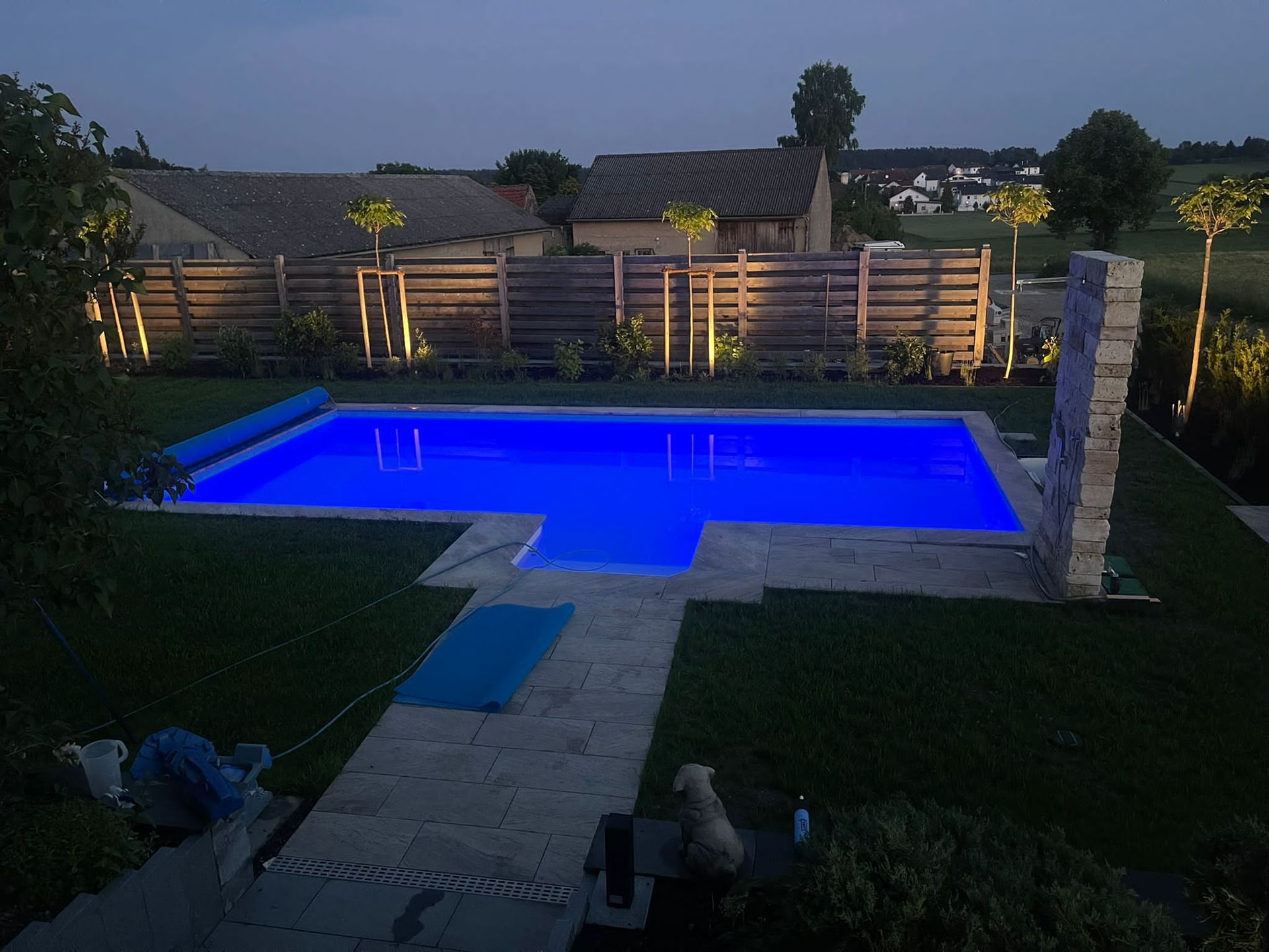 Ein in der Dämmerung blau beleuchteter Swimmingpool im Hinterhof mit Zaun und kleinem Hund.