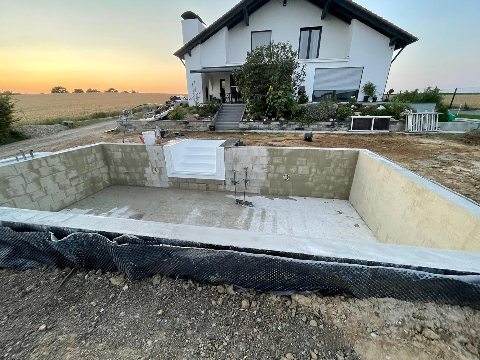 Bau eines rechteckigen Schwimmbeckens vor einem weißen Haus. Das Becken ist mit beigen Ziegeln ausgekleidet.