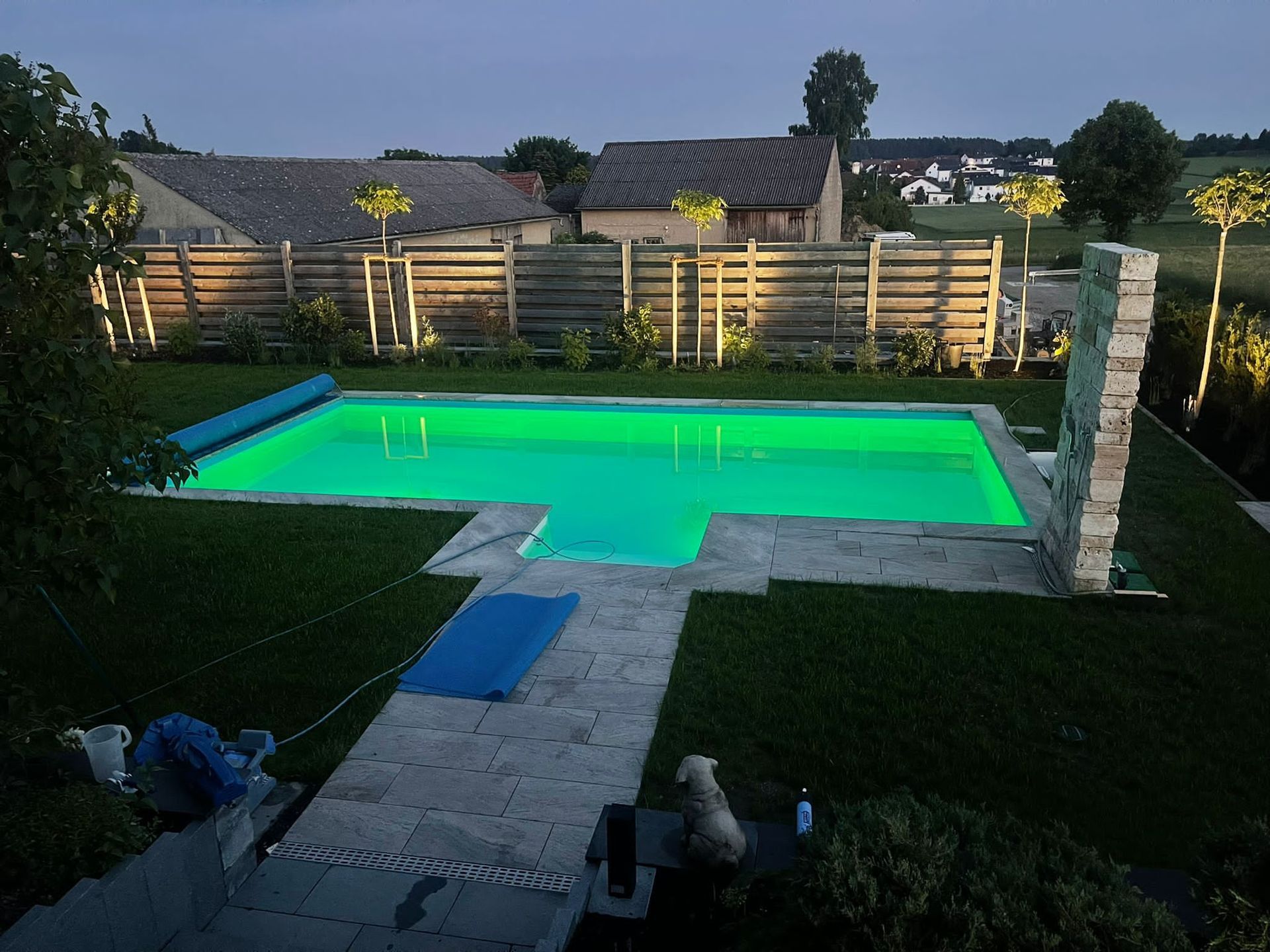 Eine Nachtansicht eines grün beleuchteten Pools, umgeben von grünem Gras, einem Steinweg und einem Holzzaun mit Lichtern.