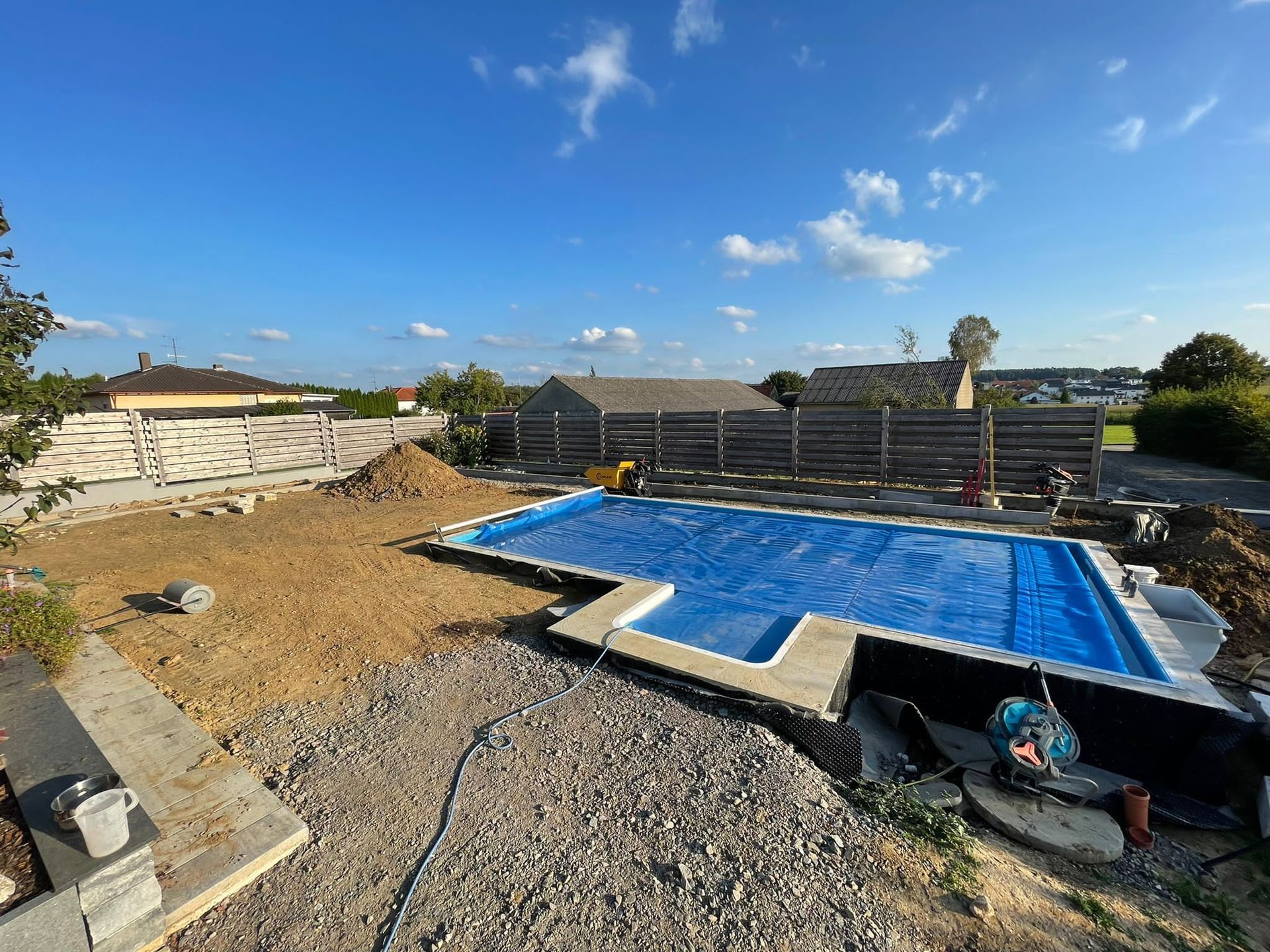 Hinterhof im Bau, rechteckiger Pool mit blauer Abdeckung, umgeben von Erde und Kies, klarer blauer Himmel.
