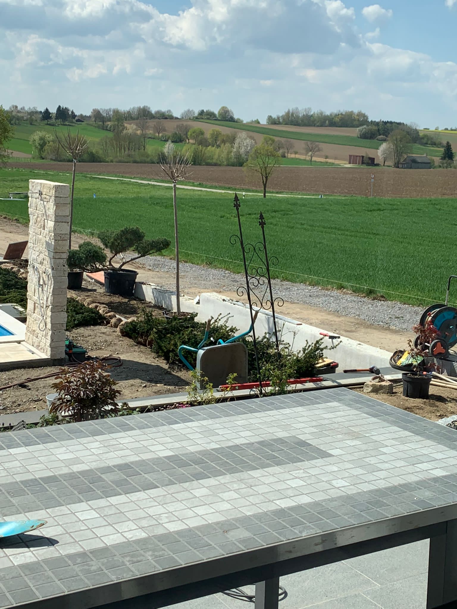Terrasse mit grauen Pflastersteinen, Landschaftsgestaltung und einem Feld im Hintergrund an einem sonnigen Tag.
