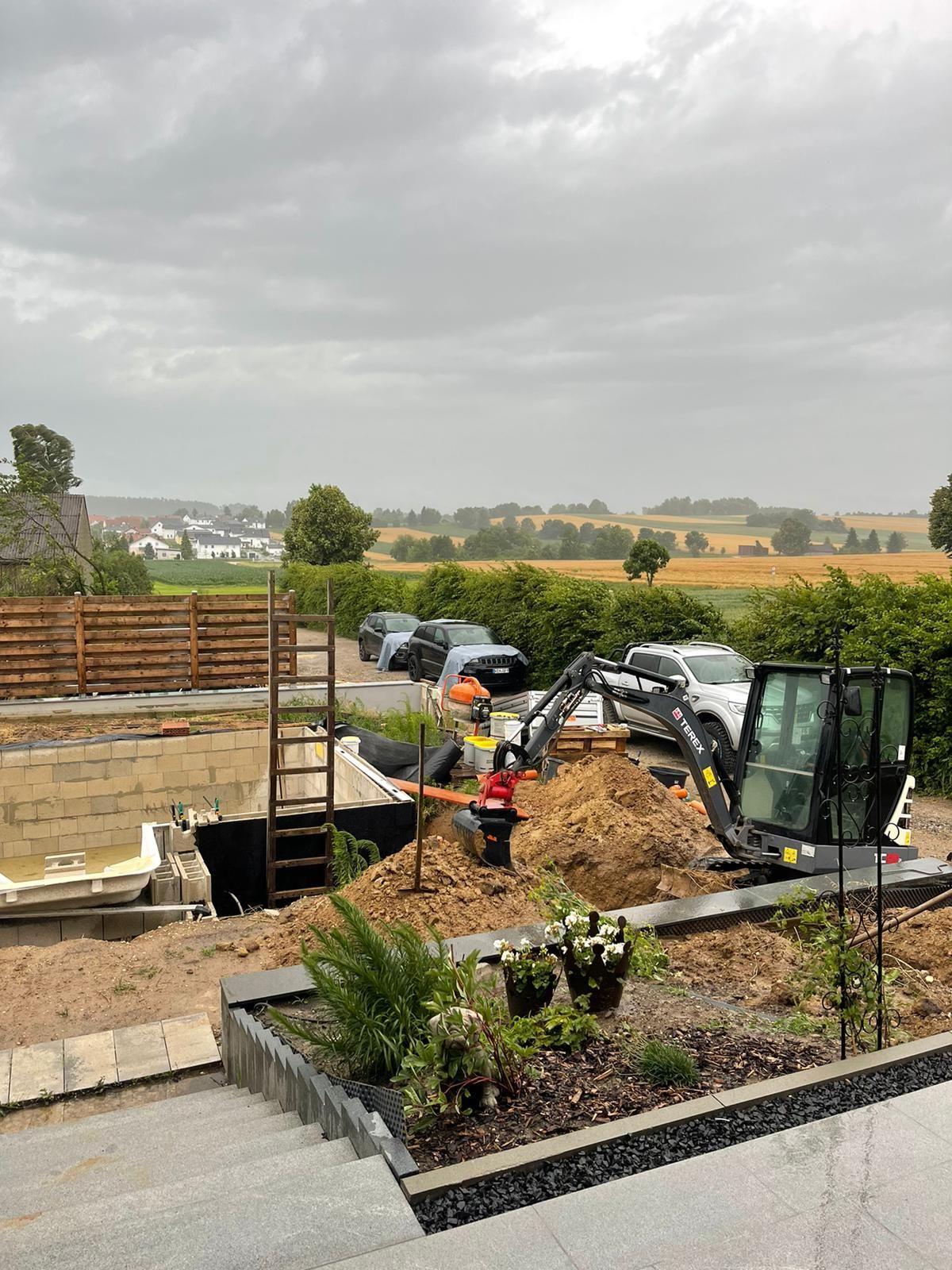 Baustelle mit Bagger, Erde, Loch und entfernter Stadt unter bewölktem Himmel.