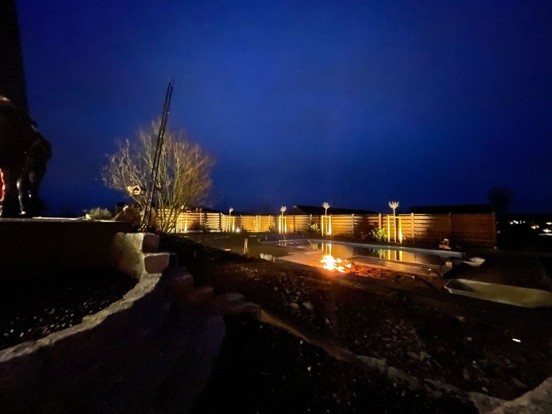Nächtliche Szene im Freien mit einem Lagerfeuer, einem Zaun und beleuchteten Bäumen unter einem dunkelblauen Himmel.