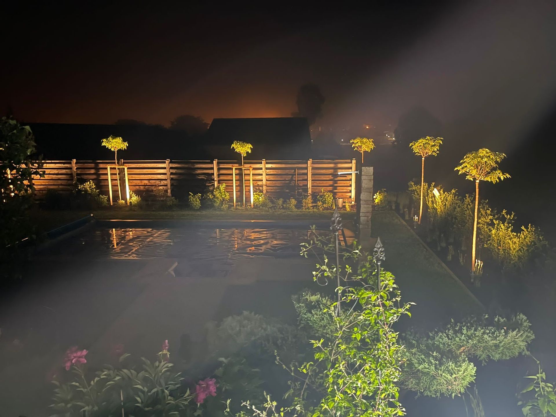 Ein nächtlicher Blick auf einen Garten mit beleuchtetem Pool, Bäumen und Zaun. Lichter leuchten und erzeugen eine neblige Atmosphäre.