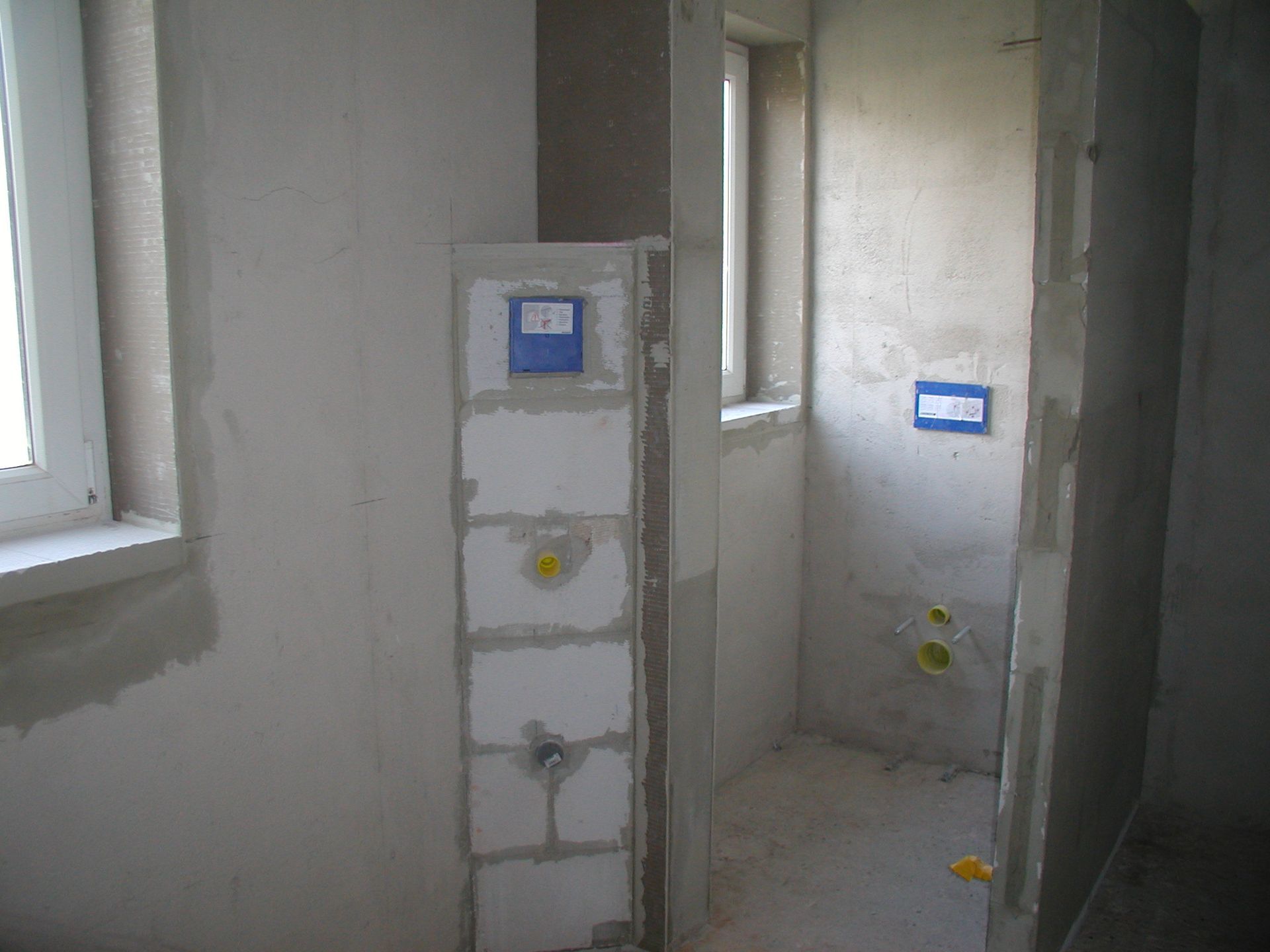 Badezimmer im Bau: weiße Wände, freiliegende Rohrleitungen, Fenster und eine Toilettenanlage aus Betonblöcken.