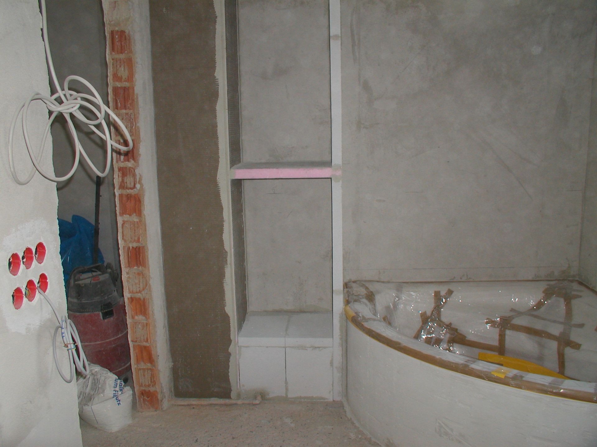 Badezimmer im Umbau; ein vertieftes Regal, eine Badewanne und freiliegende Leitungen sind sichtbar.