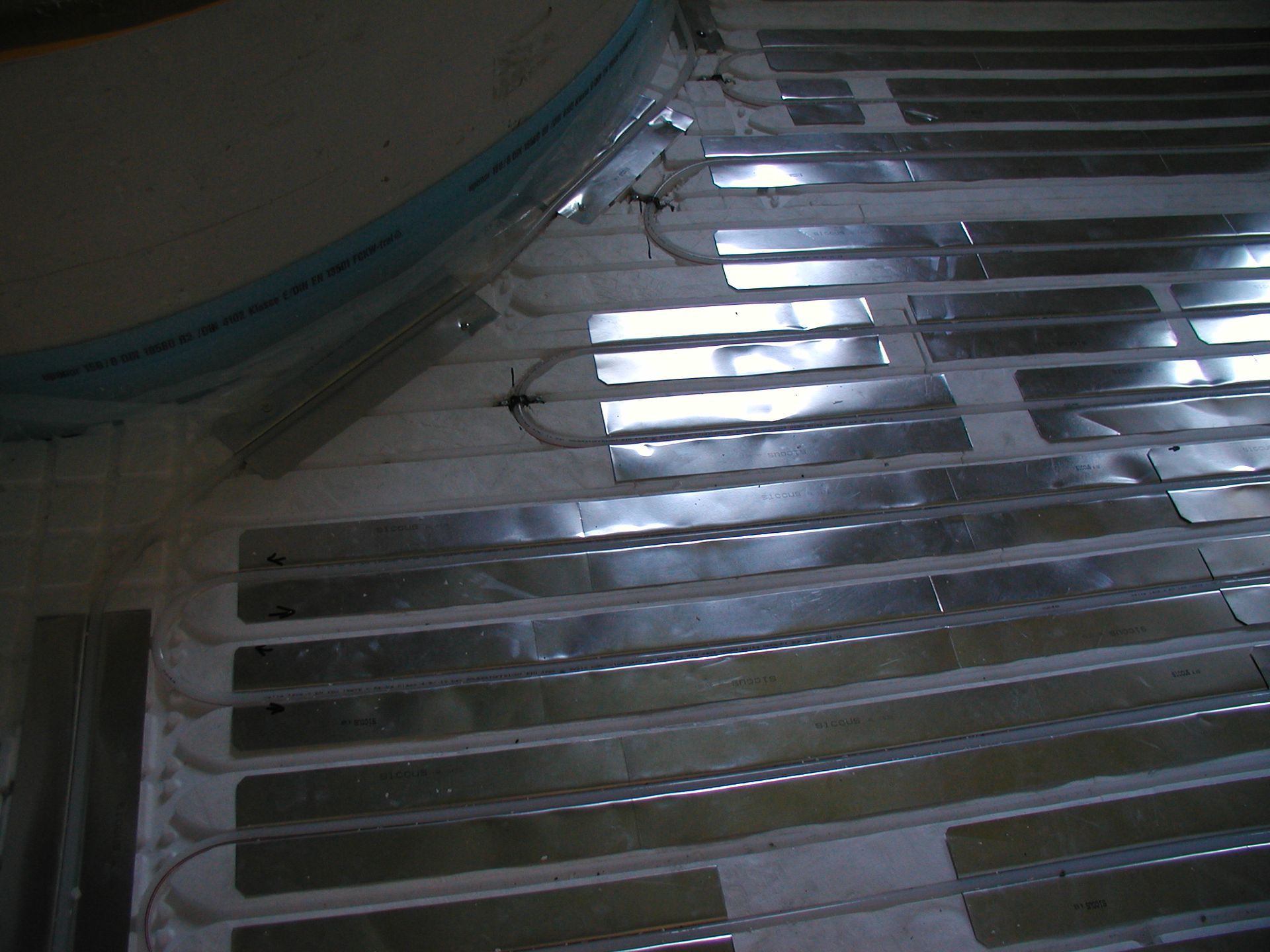 Auf einer weißen Oberfläche verlegte Aluminiumstreifen, möglicherweise für eine Fußbodenheizung im Innenbereich.