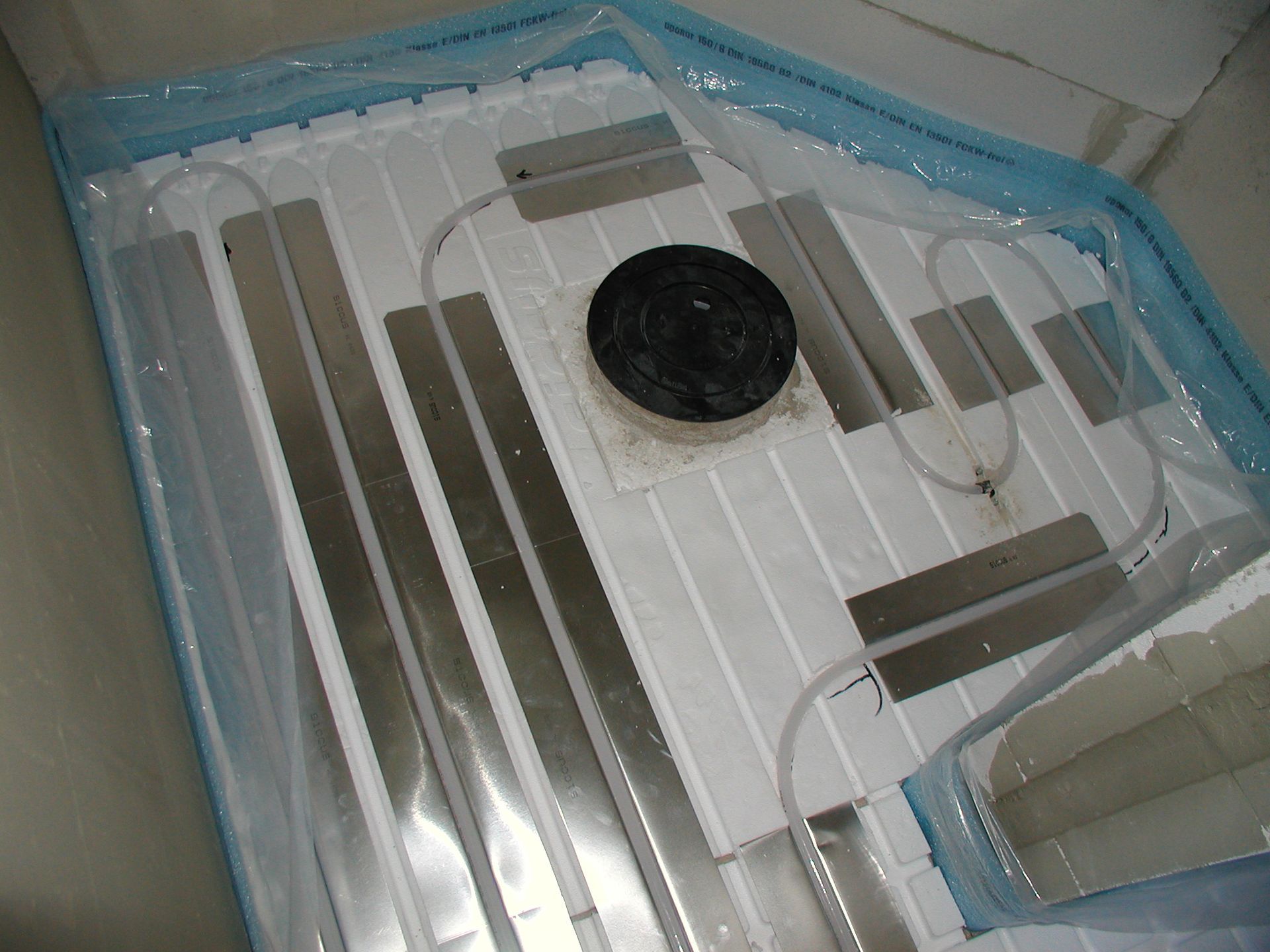 Ein Boden, der mit vakuumversiegeltem Kunststoff bedeckt ist, mit silbernen Streifen, Schläuchen und einem schwarzen runden Objekt.