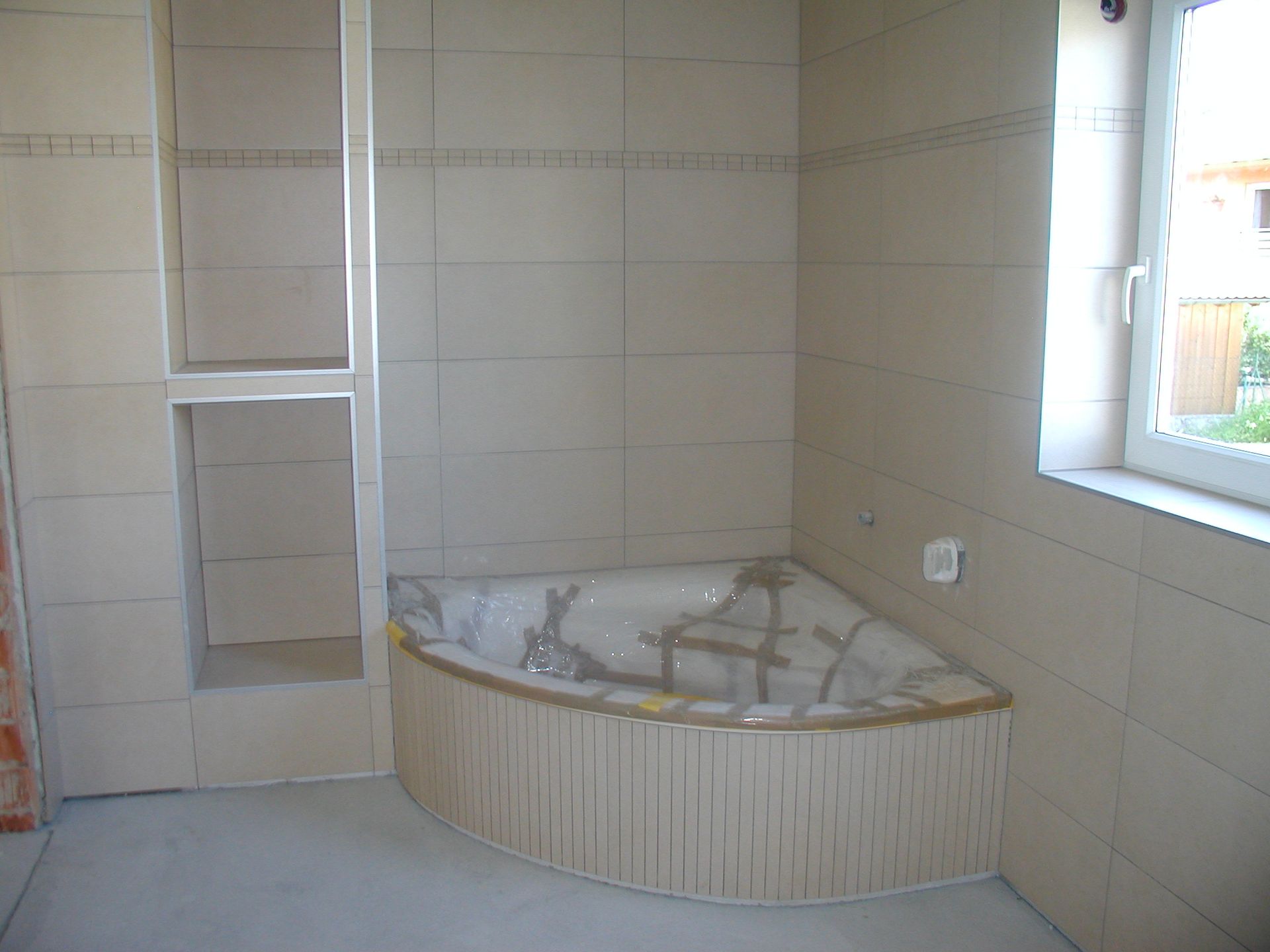 Badezimmer mit Eckbadewanne, eingebautem Regal und Fenster mit Blick ins Grüne.