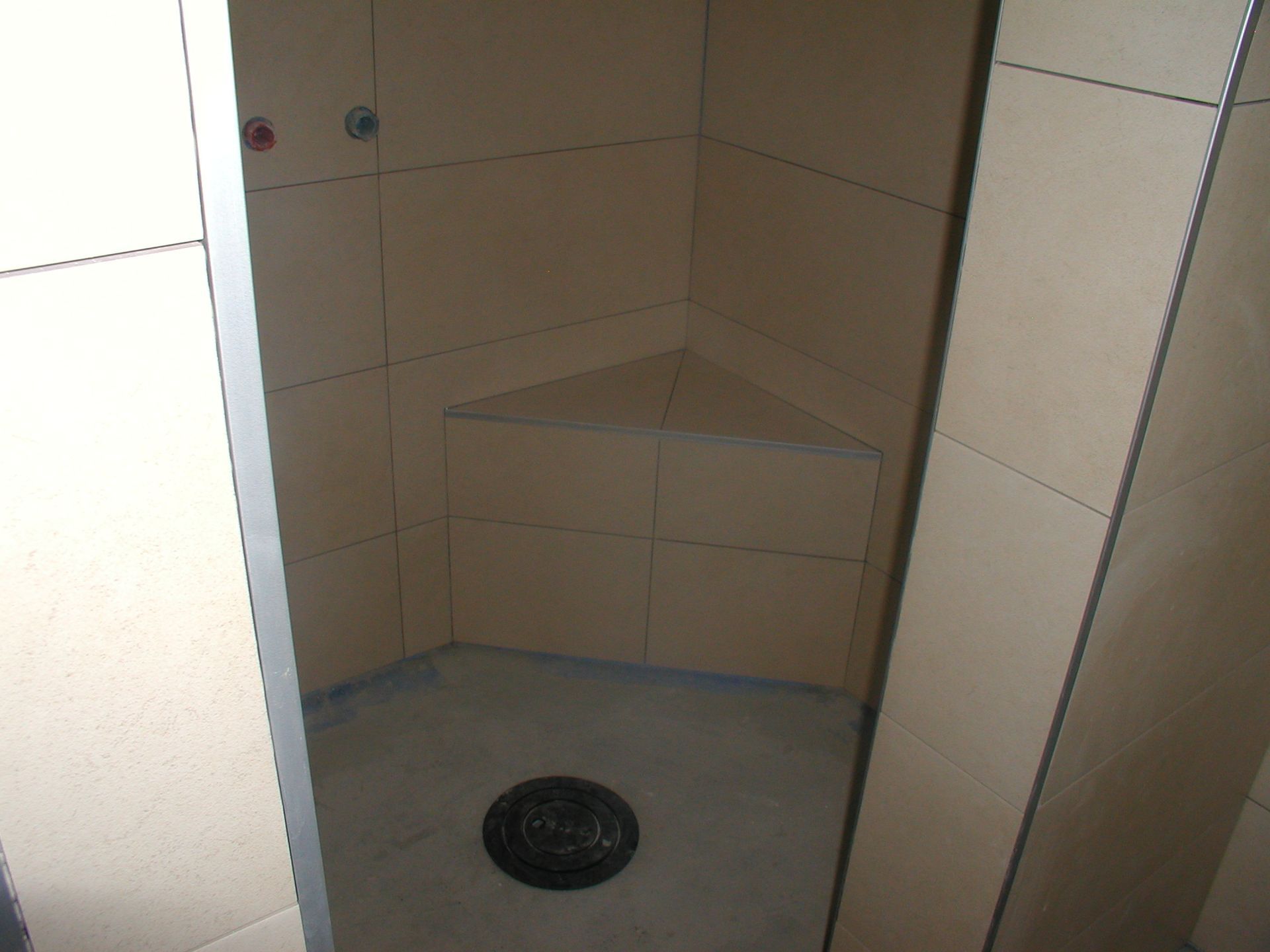 Leere Duschkabine mit eingebautem dreieckigen Sitz, hellbraunen Fliesen und Abfluss in der Mitte.