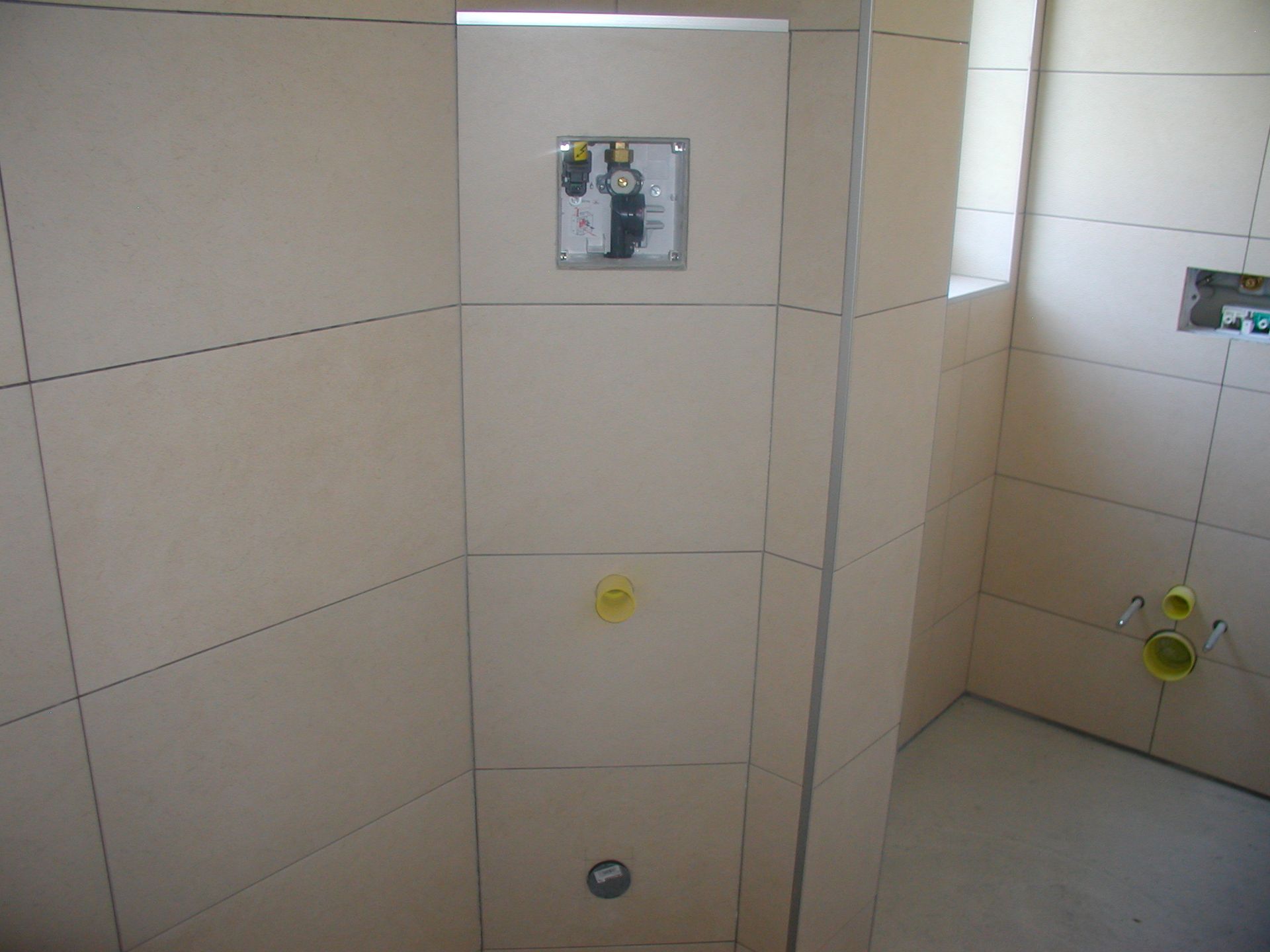 Badezimmer im Bau, beige geflieste Wände, freiliegende Rohrleitungen, Toiletten- und Waschbeckenarmaturen sichtbar.