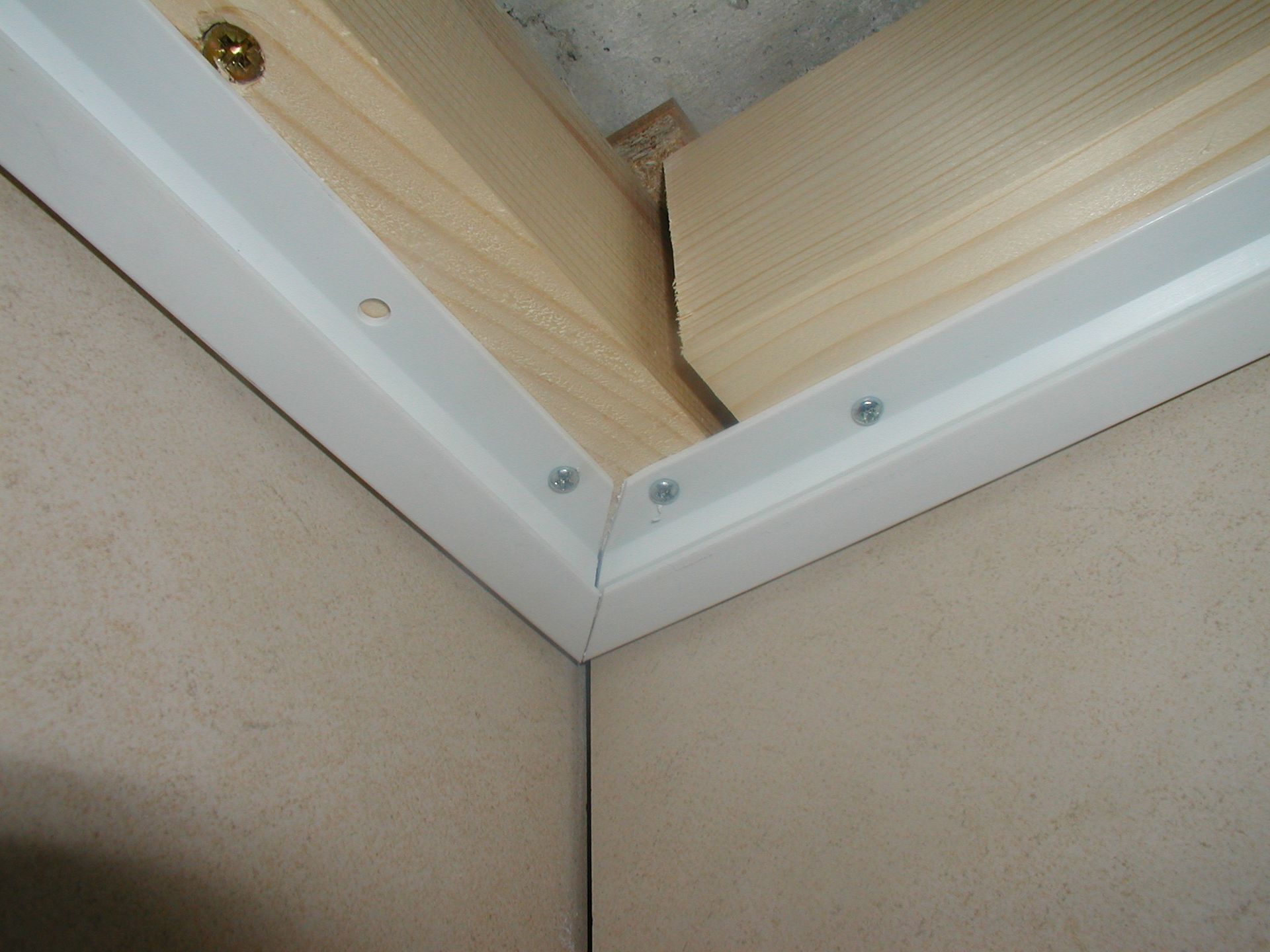 Zimmerecke mit weißen Zierleisten, Schrauben und Holzrahmen vor einer beigen Wand und Decke.