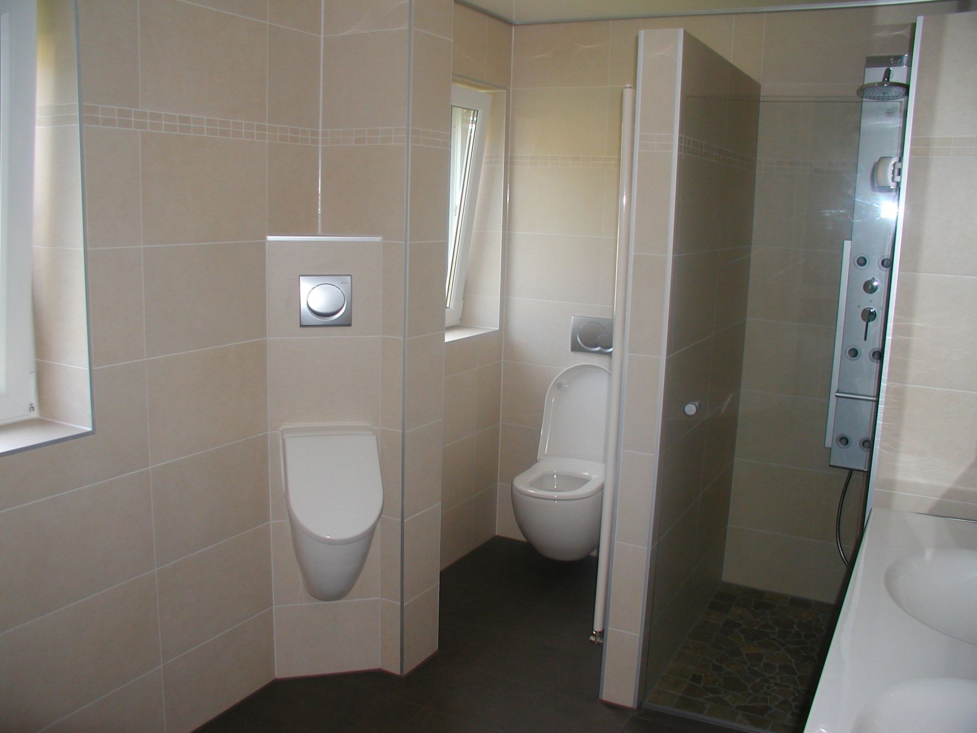 Badezimmer mit beige gefliesten Wänden, Toilette, Urinal, Duschkabine und Fenster.