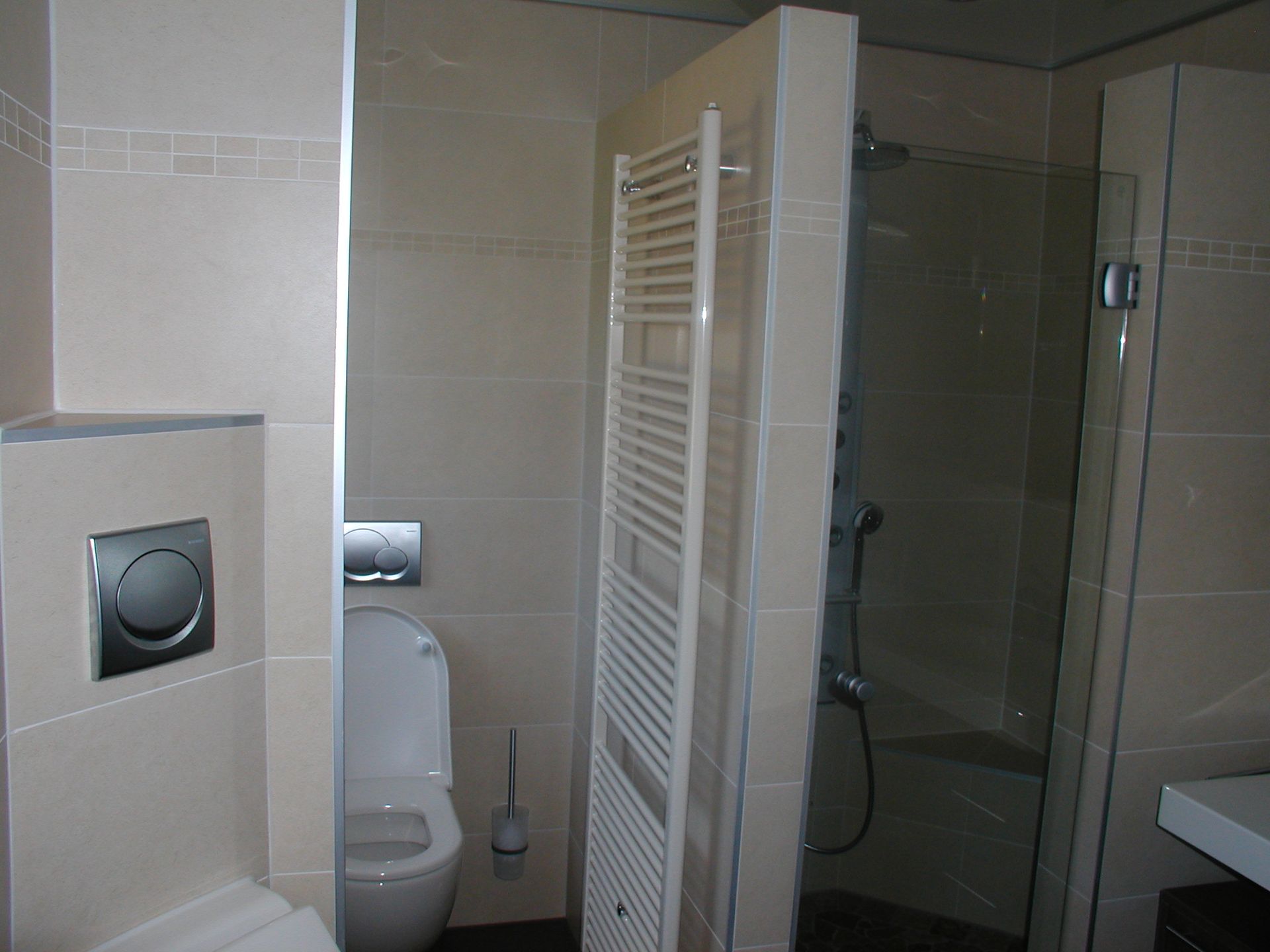 Badezimmer mit Toilette, Handtuchhalter und Dusche; beige Fliesen, Armaturen aus Edelstahl.