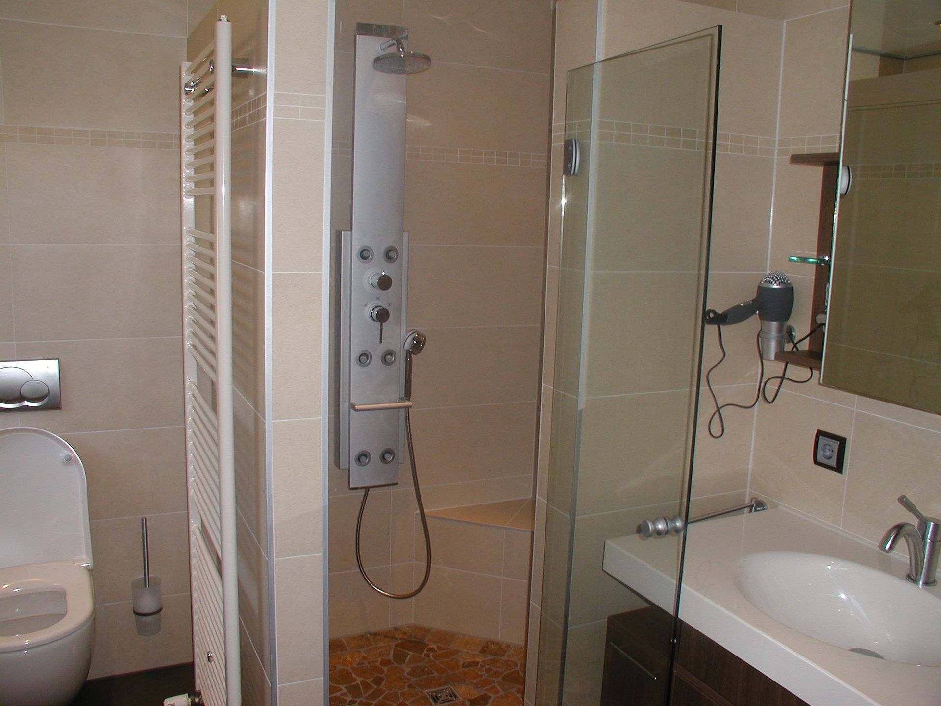 Badezimmer mit weißer Toilette, Duschpaneel, Glasduschtür, Waschbecken und Haartrockner.