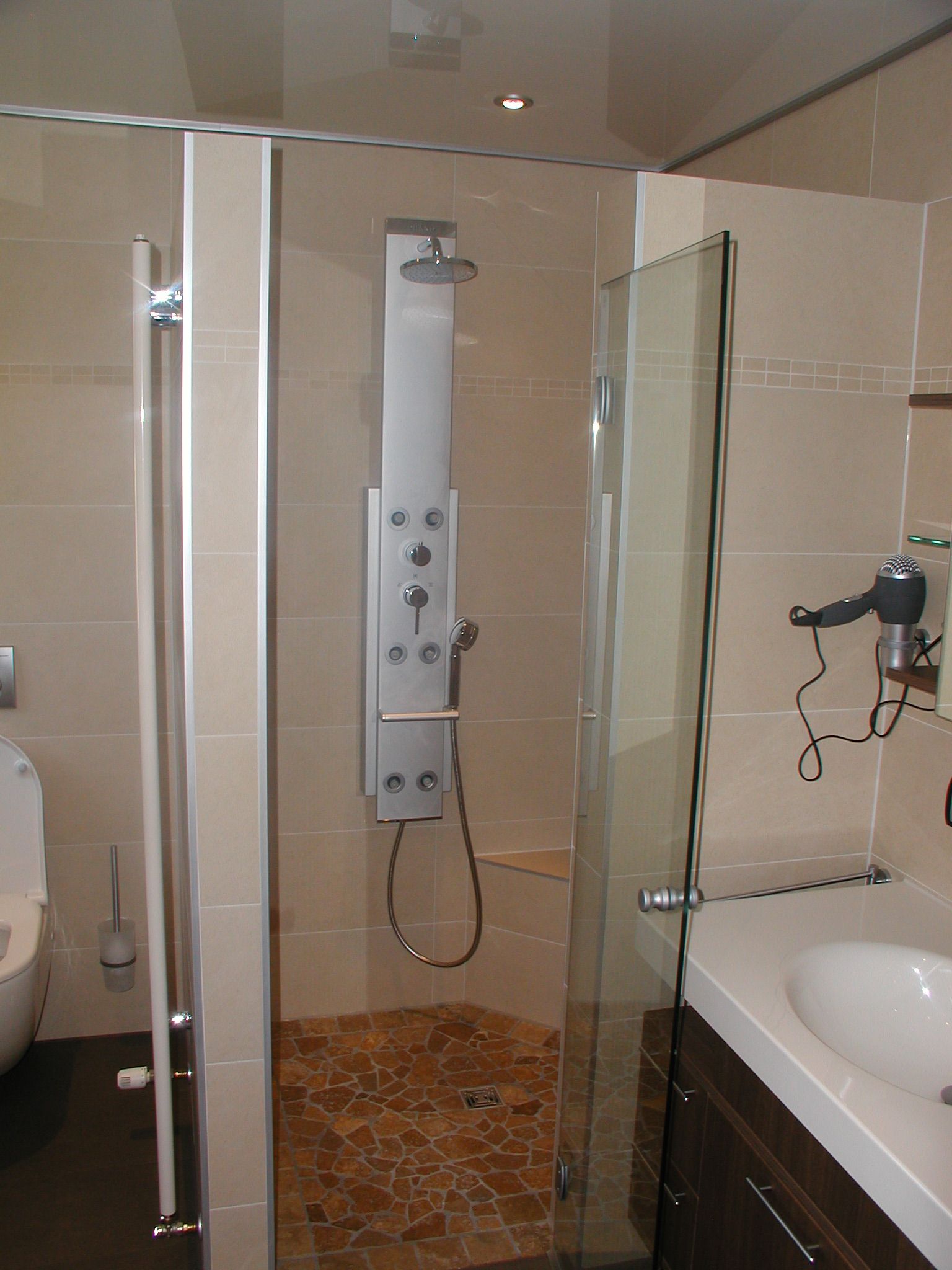 Duschkabine mit Glastüren, Steinboden, Duschpaneel und Waschbecken. Beige Wände.