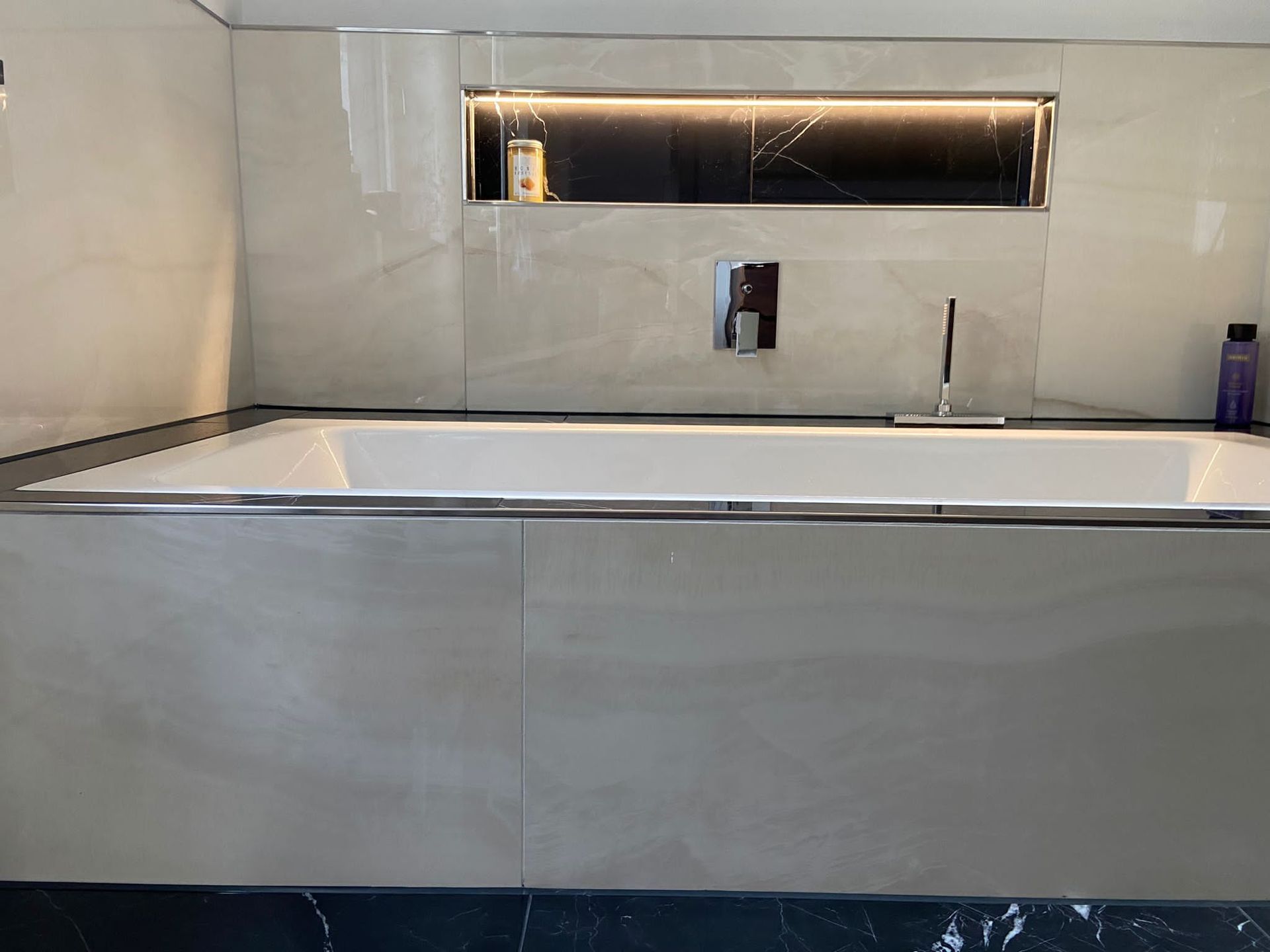 Cremefarben gefliestes Badezimmer mit eingebauter Badewanne. Edelstahlverkleidung und Wasserhahn. Eingelassenes Regal mit Flaschen über der Wanne.