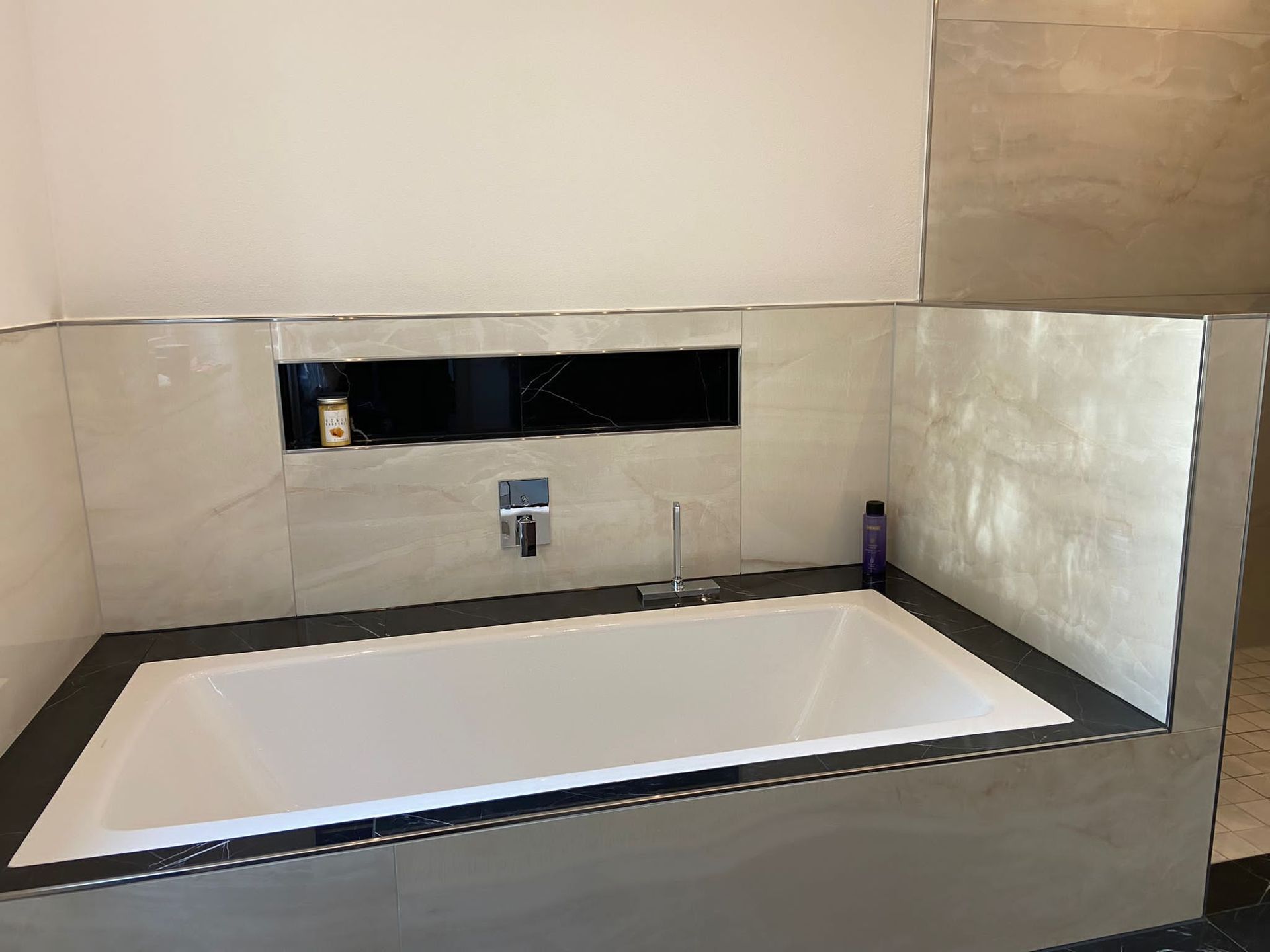 Ein modernes Badezimmer mit eingebauter Badewanne, gefliesten Wänden und einem Regal mit Toilettenartikeln.