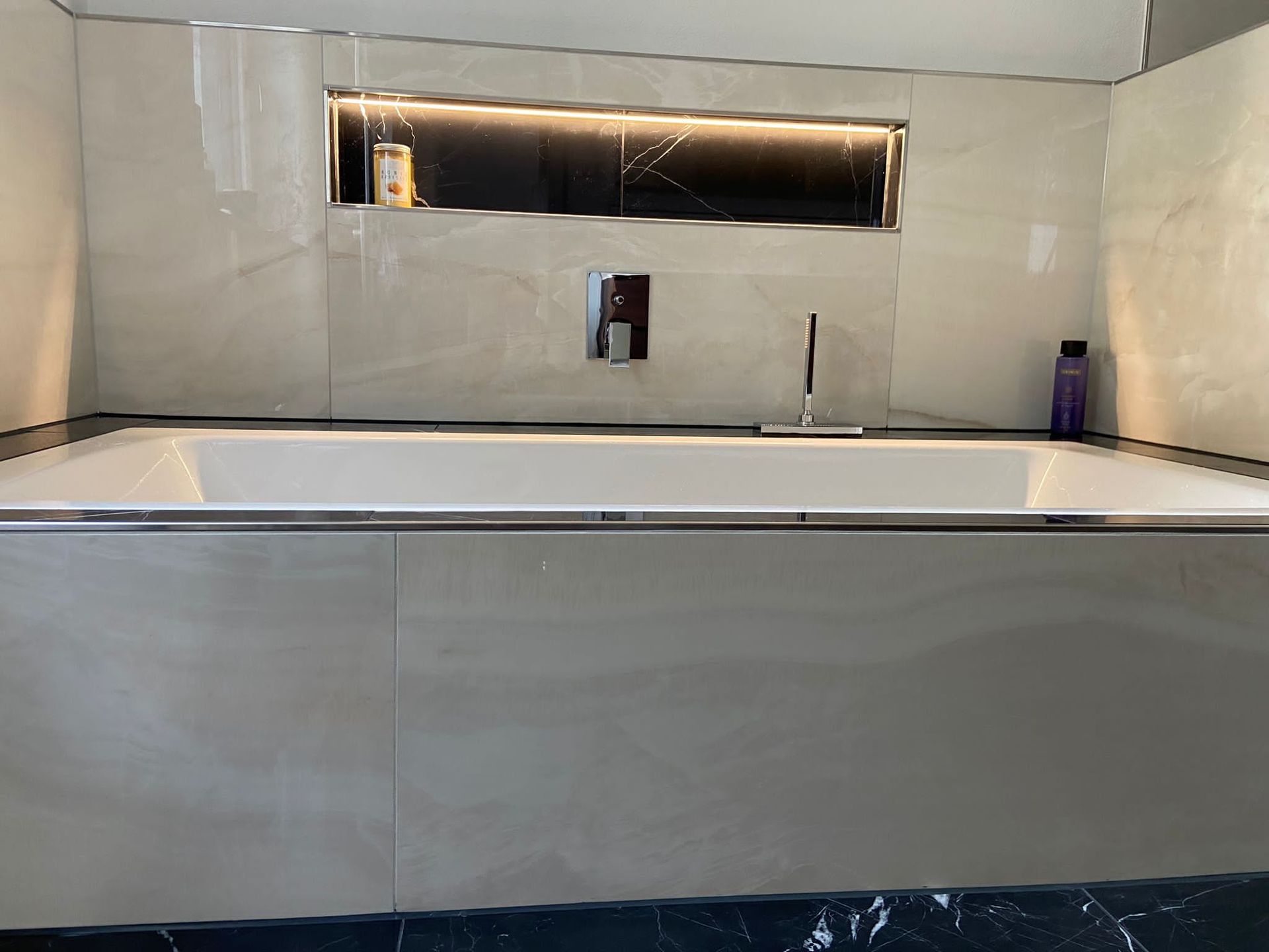 Modernes Badezimmer mit gefliester Badewanne, Chromhahn und einem Einbauregal aus dunklem Marmor.