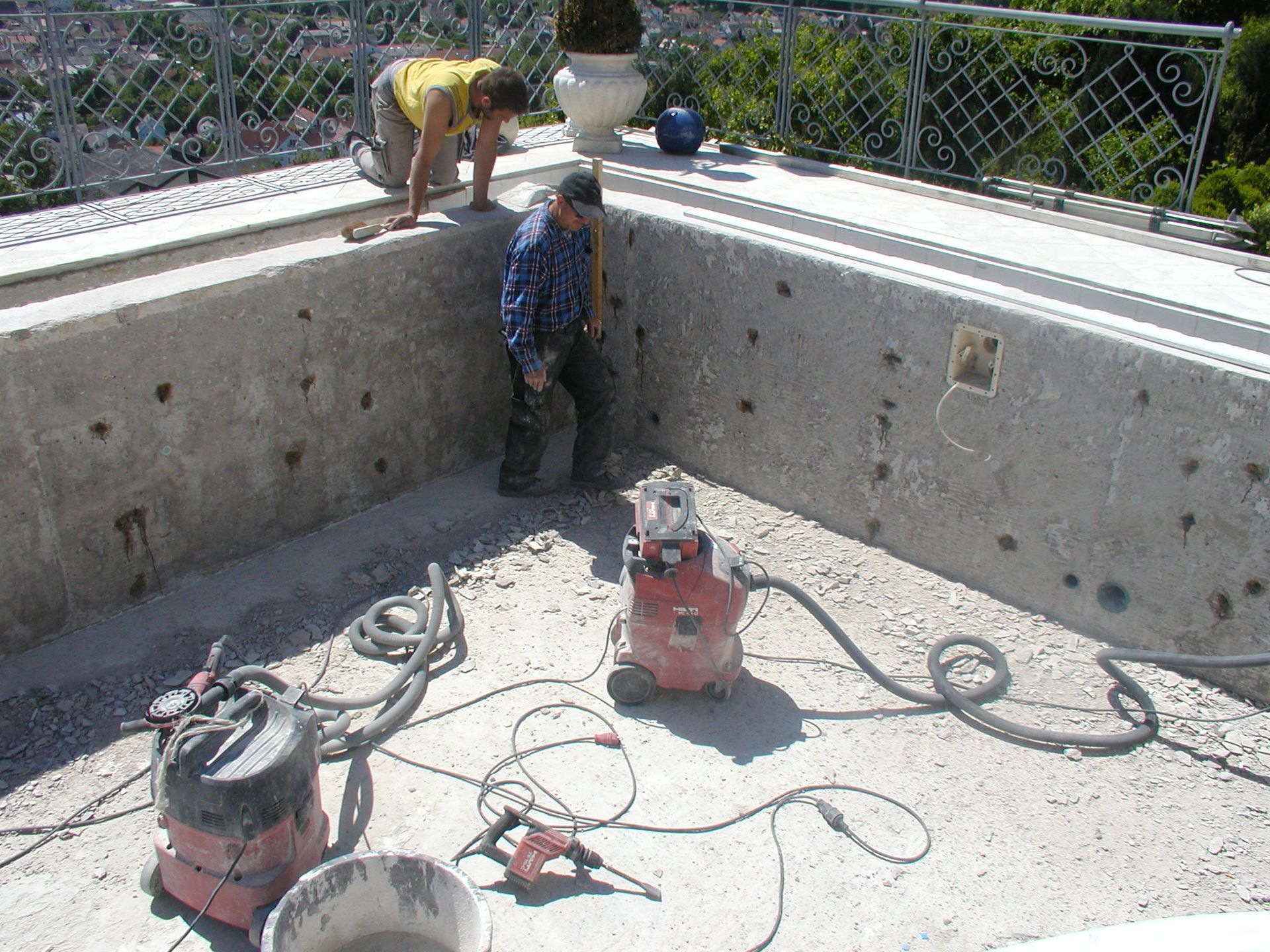 Zwei Arbeiter renovieren ein Betonbecken; einer kniet, der andere steht. Rote Maschine, Werkzeuge auf dem Boden. Dachterrasse.