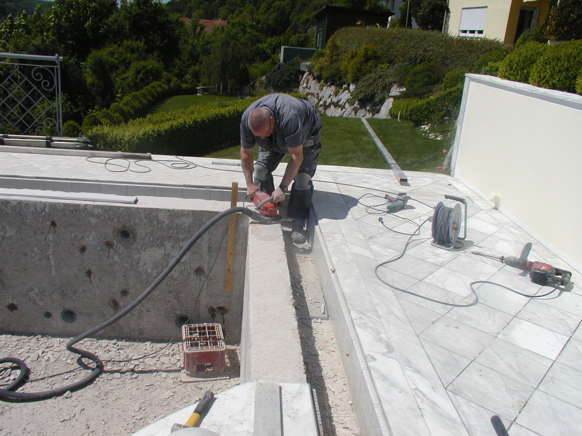 Mann schneidet mit einer Säge Steine auf einer Terrasse, umgeben von Baumaterialien und einem üppigen grünen Hintergrund.