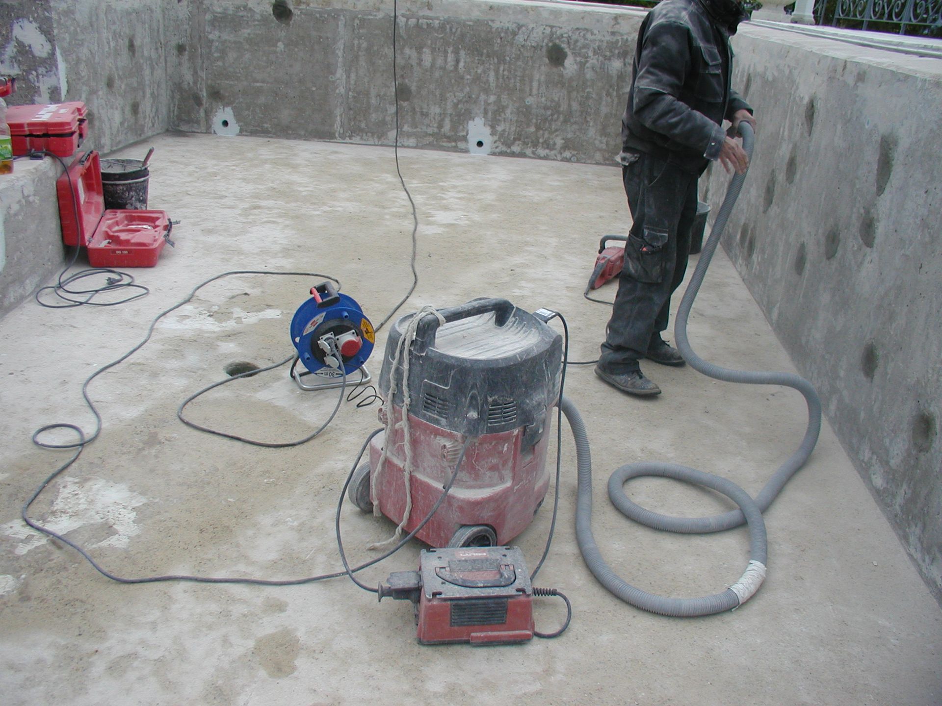 Ein Arbeiter saugt eine Betonoberfläche ab, Staub und Geräte sind in dem unfertigen Raum verstreut.