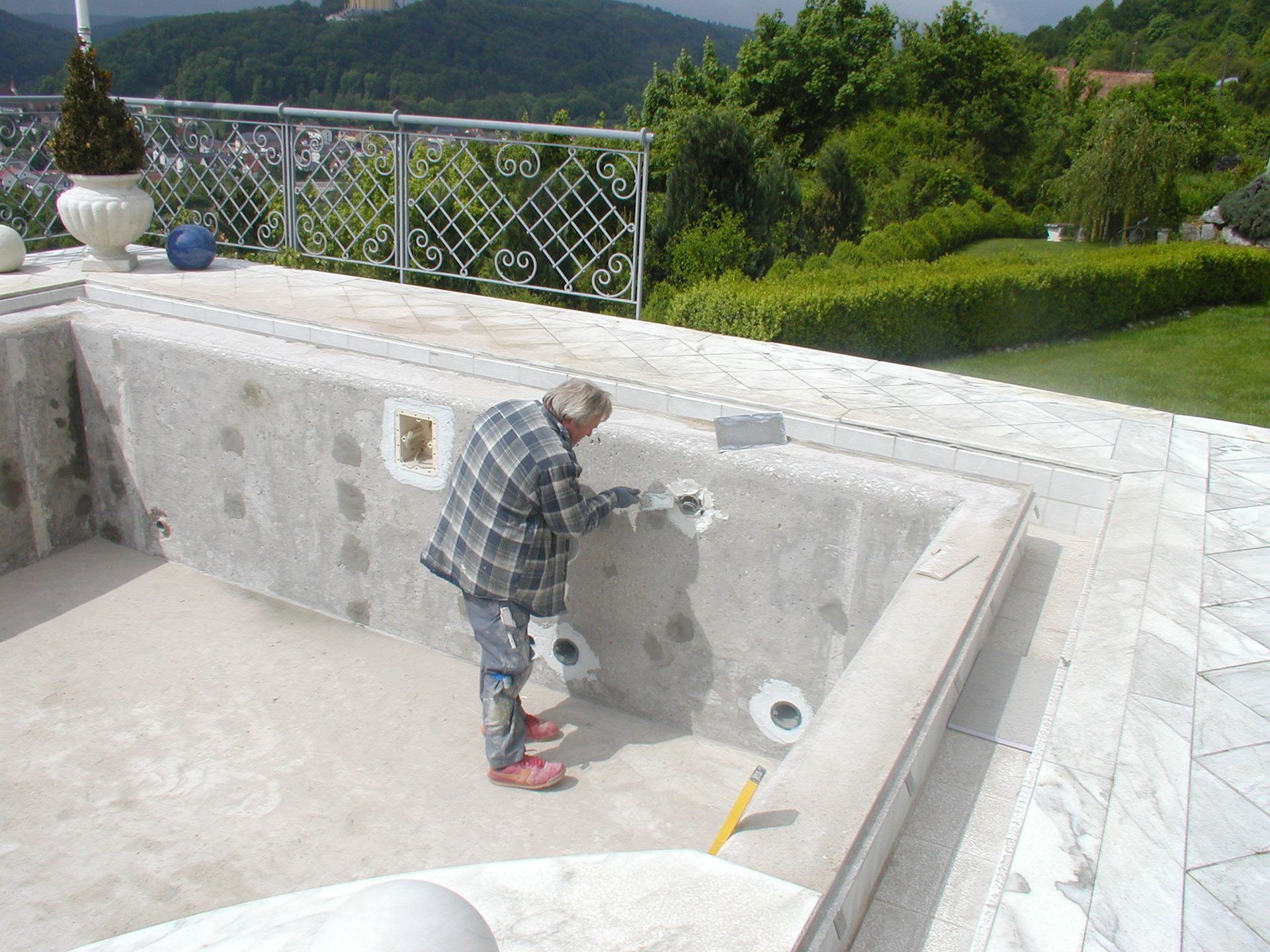 Mann im karierten Hemd repariert einen leeren Swimmingpool mit Berghintergrund.