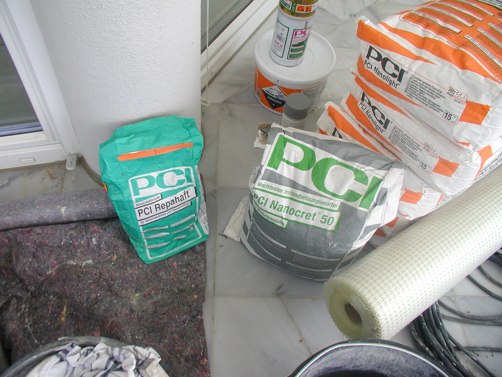 Taschen mit PCI-Baumaterialien, darunter grüne und orangefarbene Taschen, mit anderen Werkzeugen auf einer Marmoroberfläche.