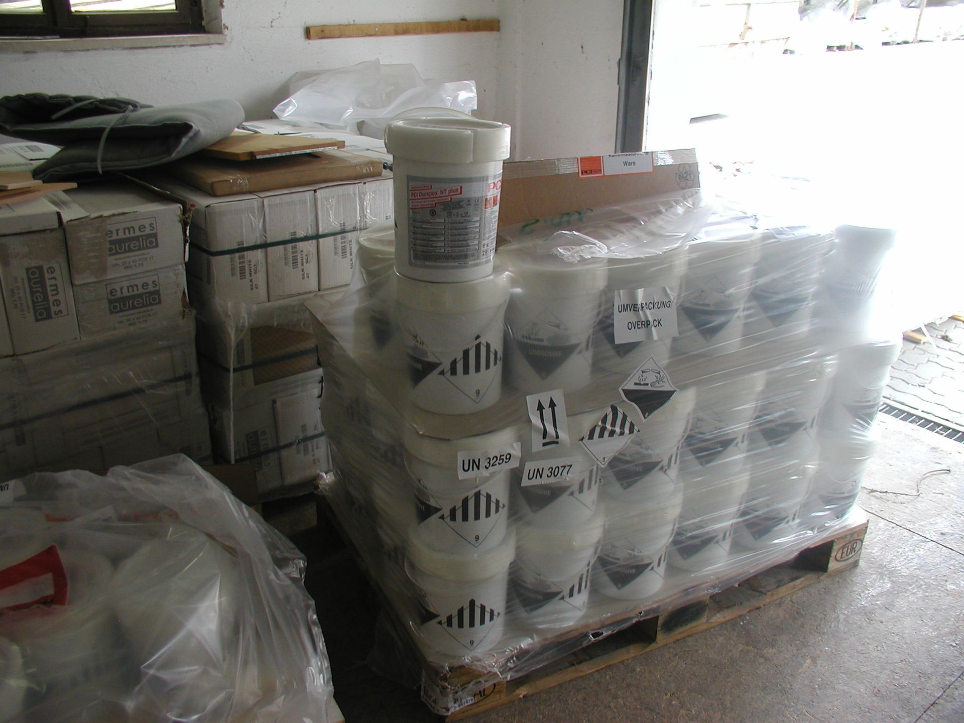 Eine Palette mit weißen Chemikalieneimern, in durchsichtige Plastikfolie eingewickelt, gestapelt in einem Lagerhaus. Gefahrstoffsymbole sichtbar.
