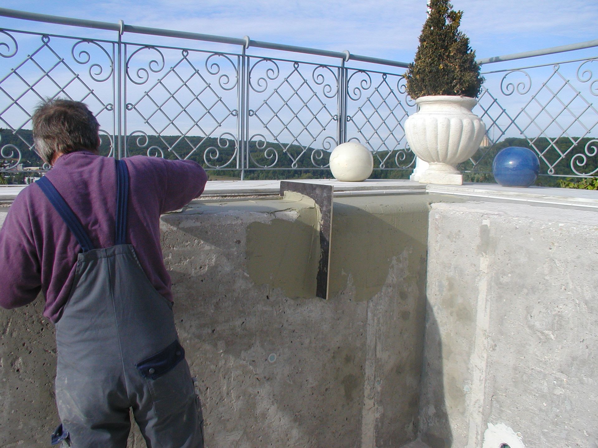 Mann im Overall trägt Mörtel auf eine Betonwand auf, im Hintergrund verziertes Geländer und Topfpflanze.