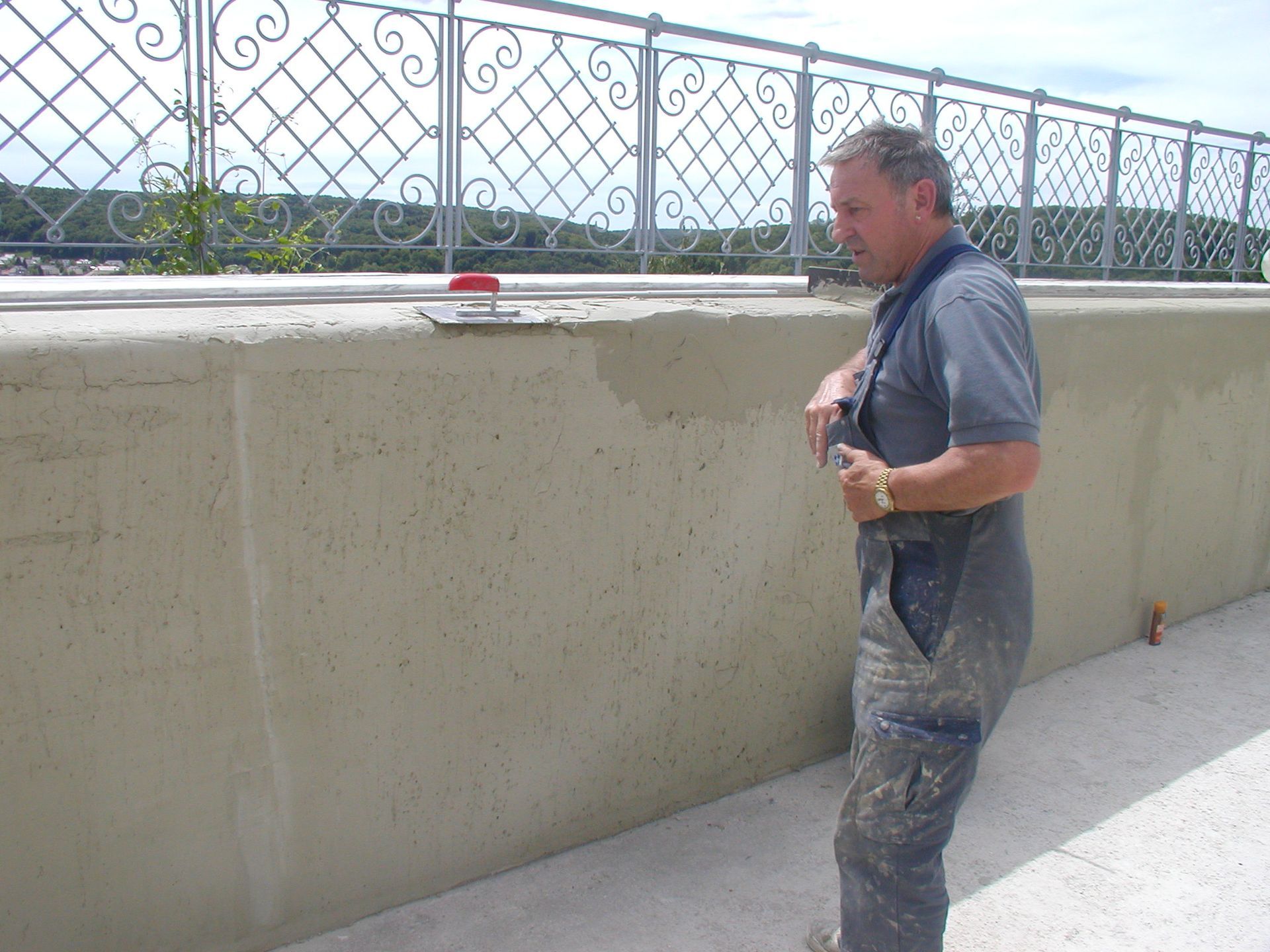 Mann verputzt im Freien eine helle Wand neben einem dekorativen Geländer.
