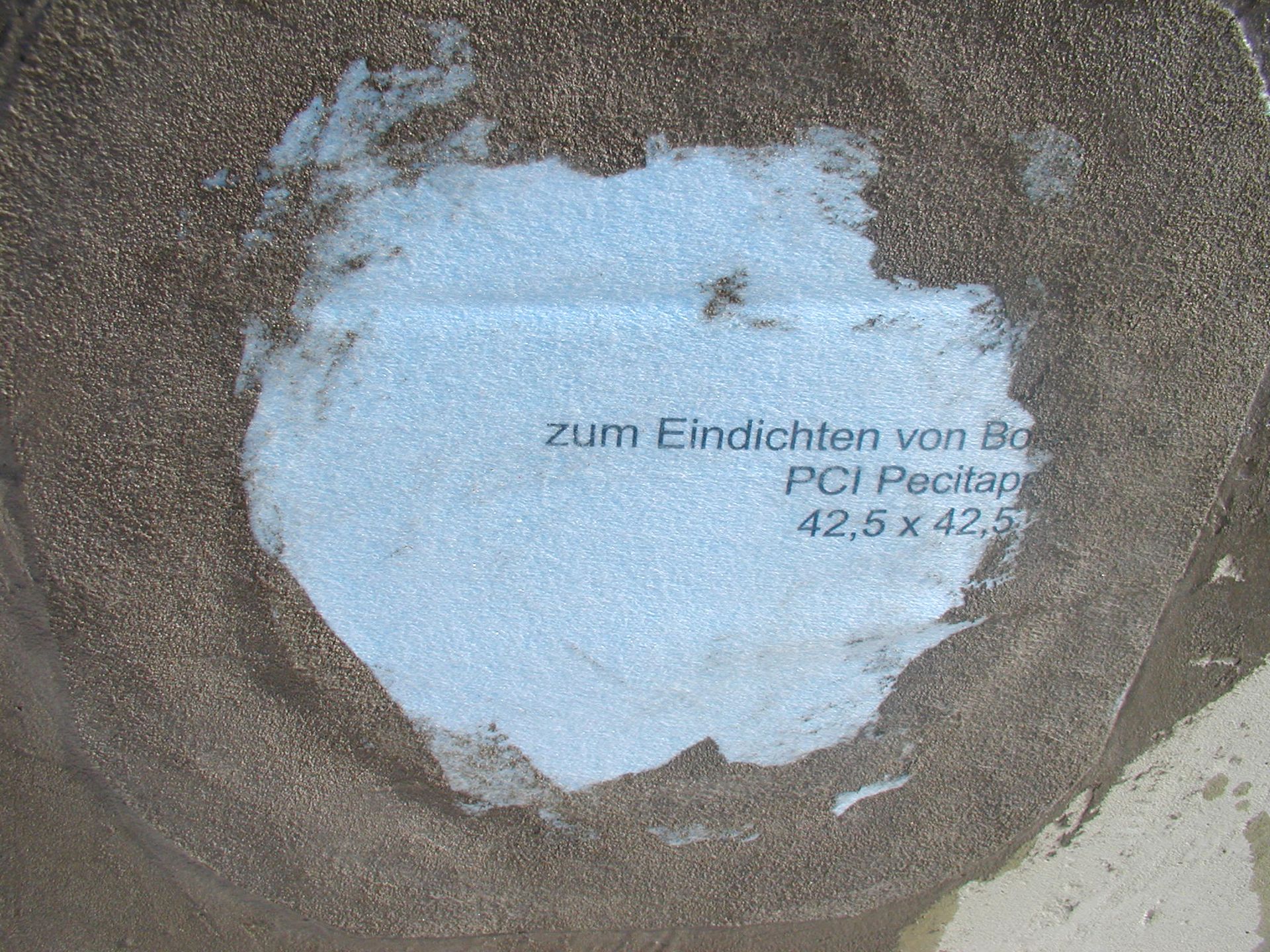 Text auf einem hellblauen quadratischen Flicken, der auf eine strukturierte braune Oberfläche geklebt ist; lautet „Zum Eindichten von Ba...“ und „PCI Pecitap 42,5 x 42,5.“