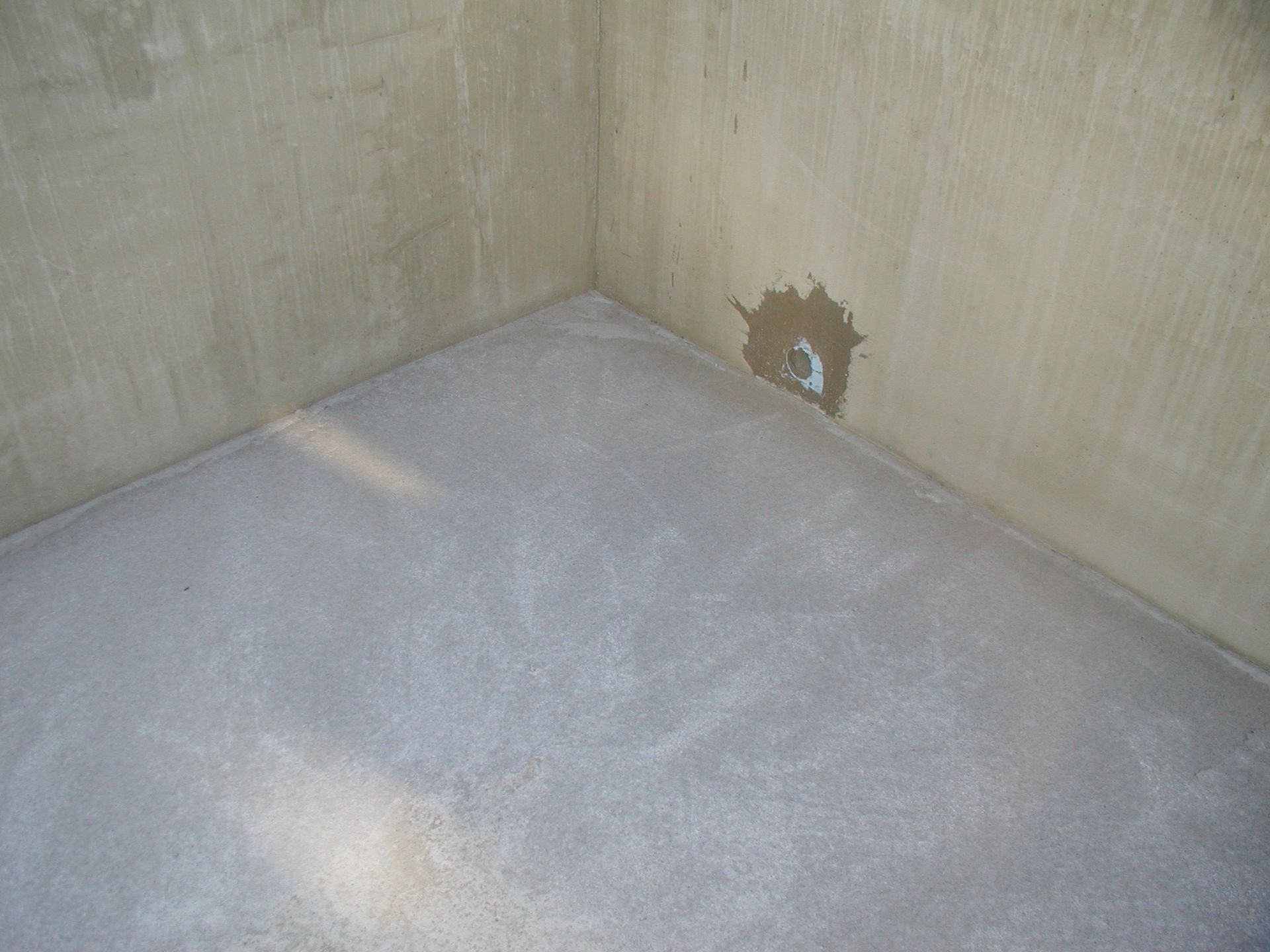Ecke eines Raumes mit grauem Boden und hellbraunen Wänden; ein Teil der Wand ist beschädigt.