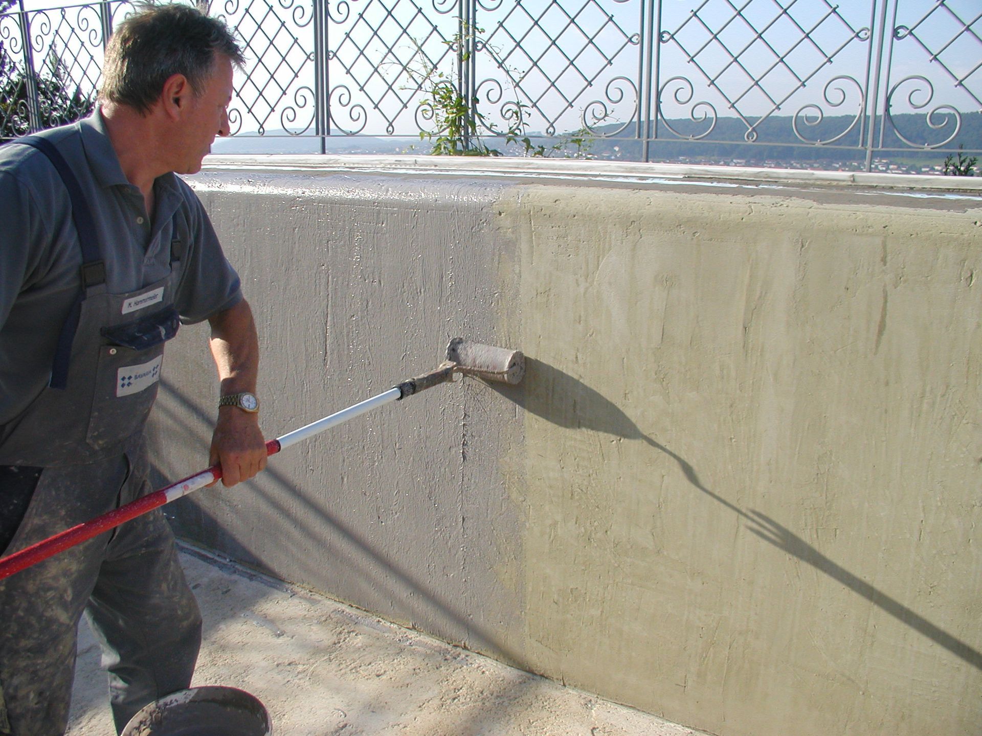 Mann streicht eine Betonwand mit einer Rolle. Er ist an einem sonnigen Tag draußen und streicht mit einer Rolle mit langem Griff.