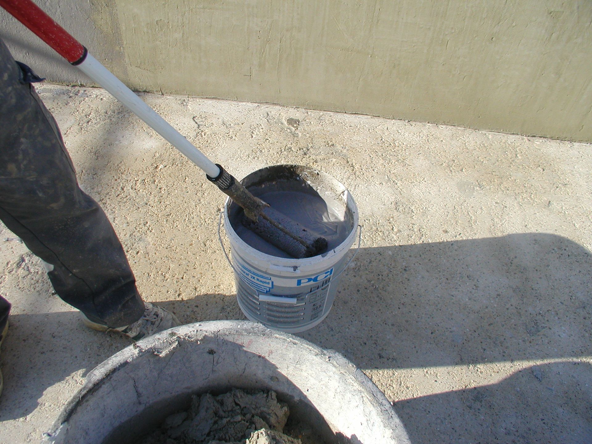 Person, die mit einer Farbrolle eine graue Flüssigkeit aus einem Eimer auf eine Betonoberfläche aufträgt.