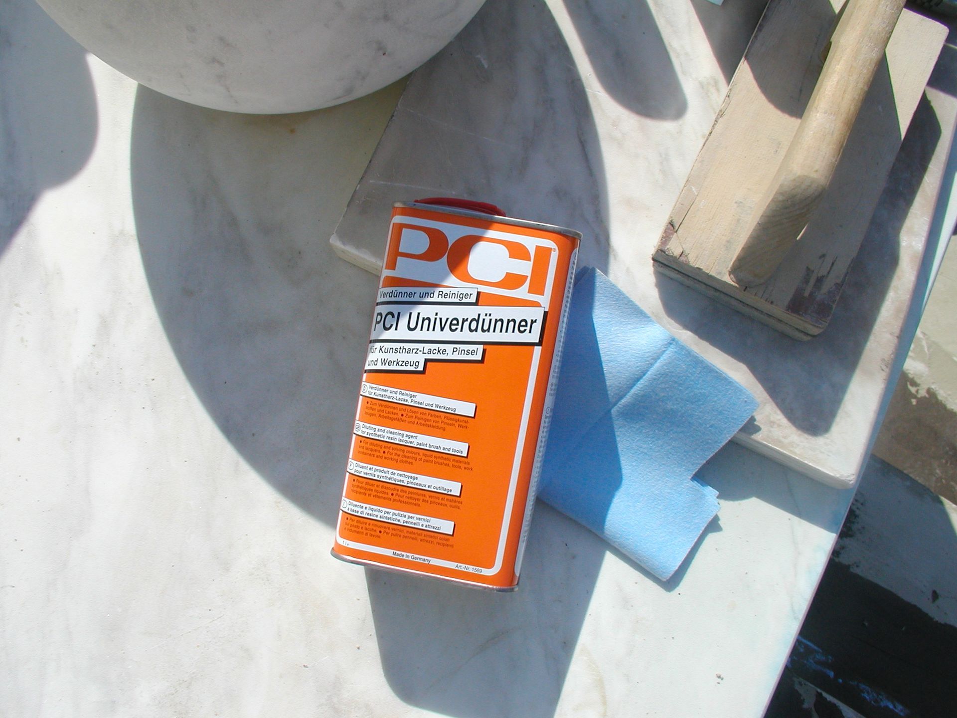 Orangefarbene PCI-Lösungsmitteldose, blaues Tuch und Holzwerkzeuge auf einer Marmoroberfläche.